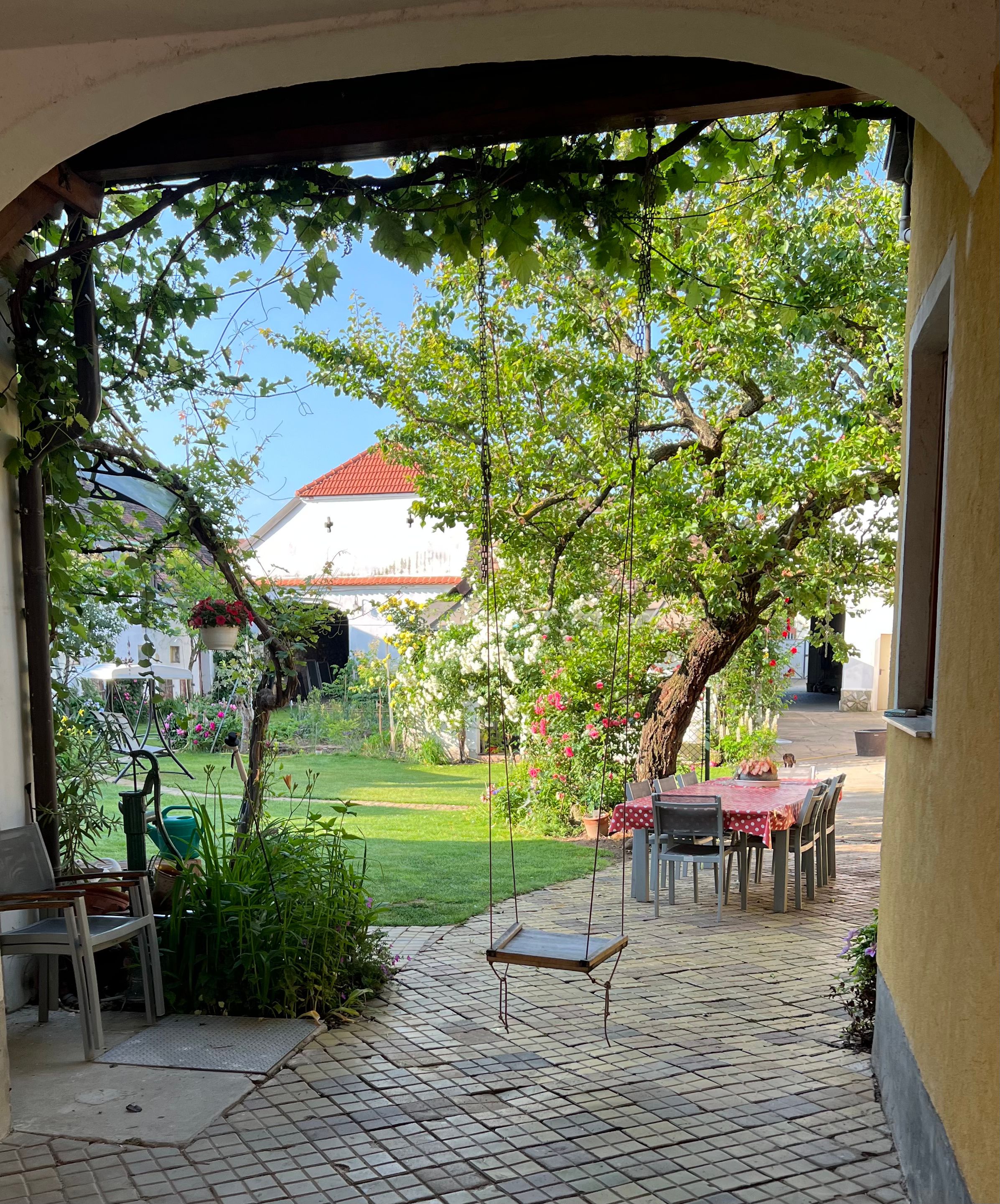 Idylická záhrada s hojdačkou, stolom so stoličkami a kvitnúcimi rastlinami.