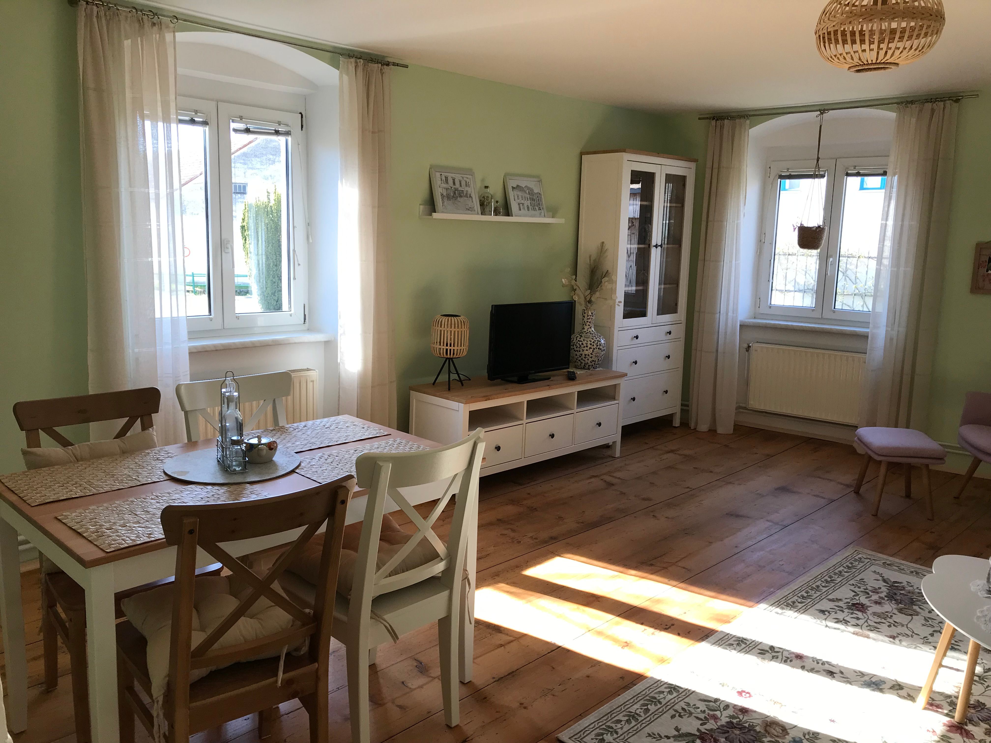 Svetlá izba s jedálenským stolom, stoličkami, TV skrinkou a veľkými oknami.