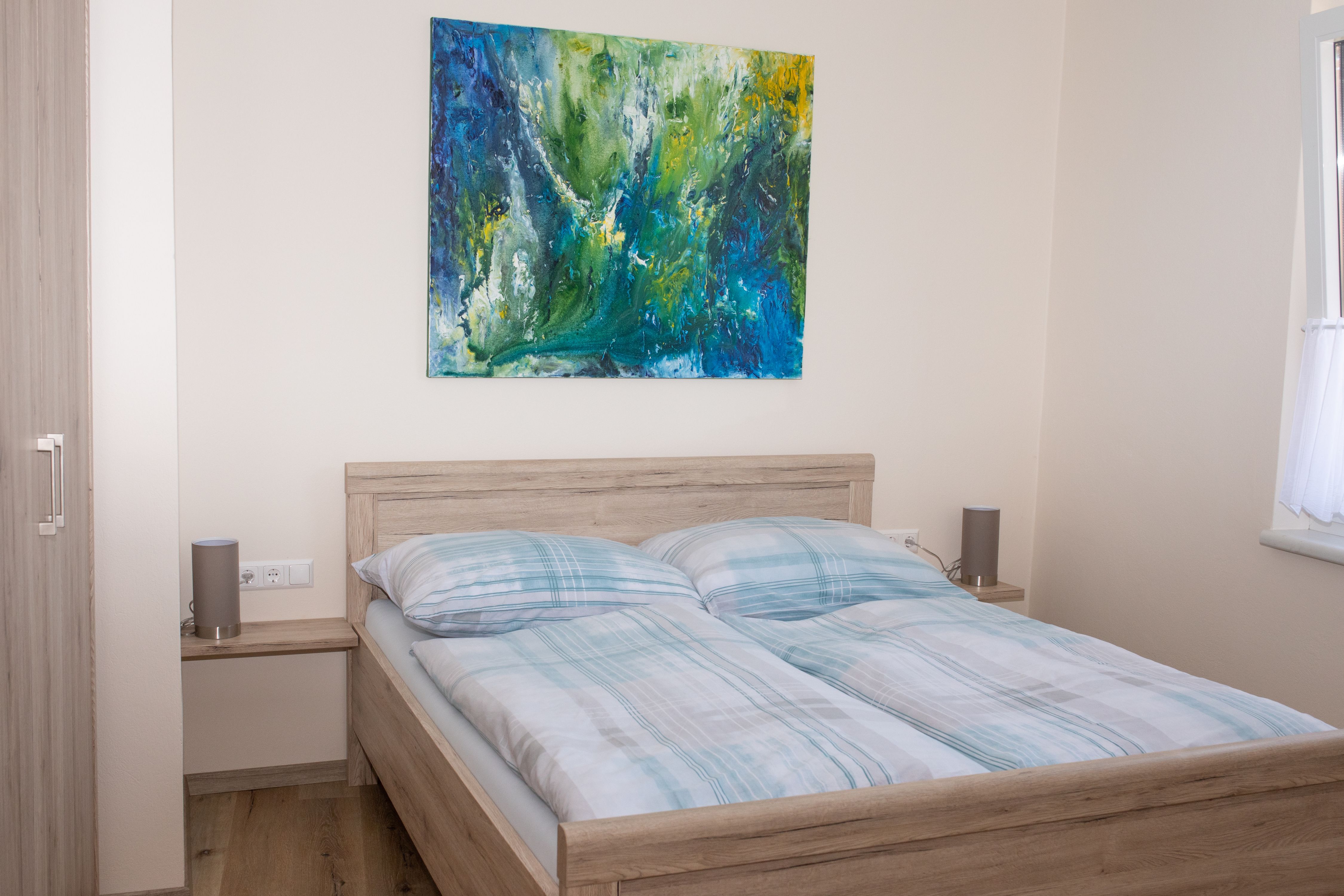 Spálňa s manželskou posteľou, abstraktnou maľbou na stene a oknom so závesom.