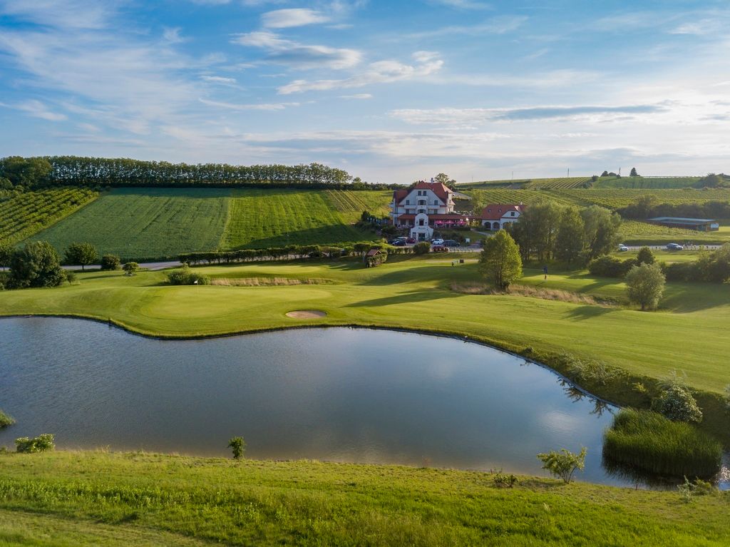 Záber na golfové ihrisko s rybníkom a klubovňou v pozadí.