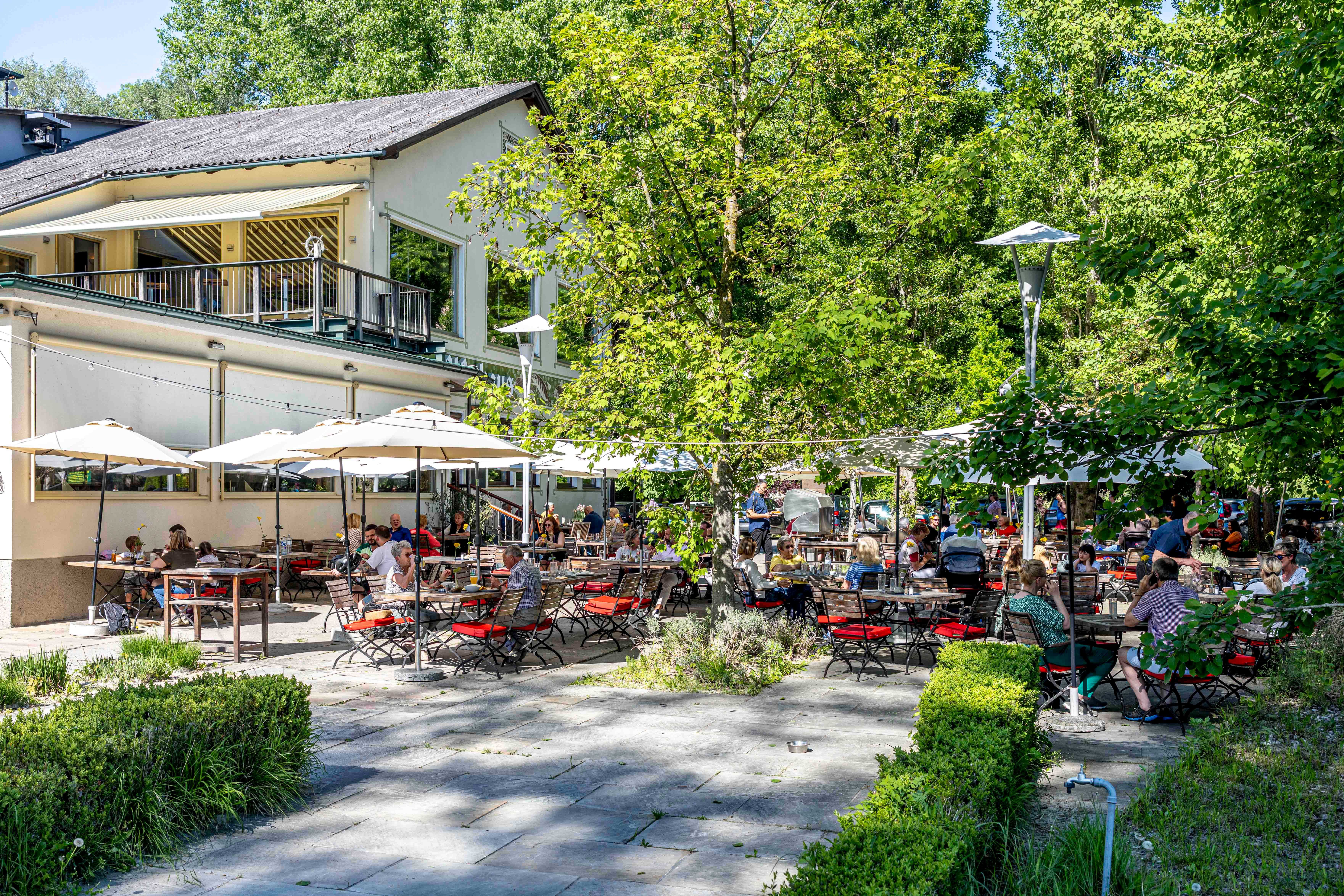 Záhrada pre hostí s množstvom ľudí sediacich pri stoloch pod slnečníkmi, obklopená stromami a budovou v pozadí.