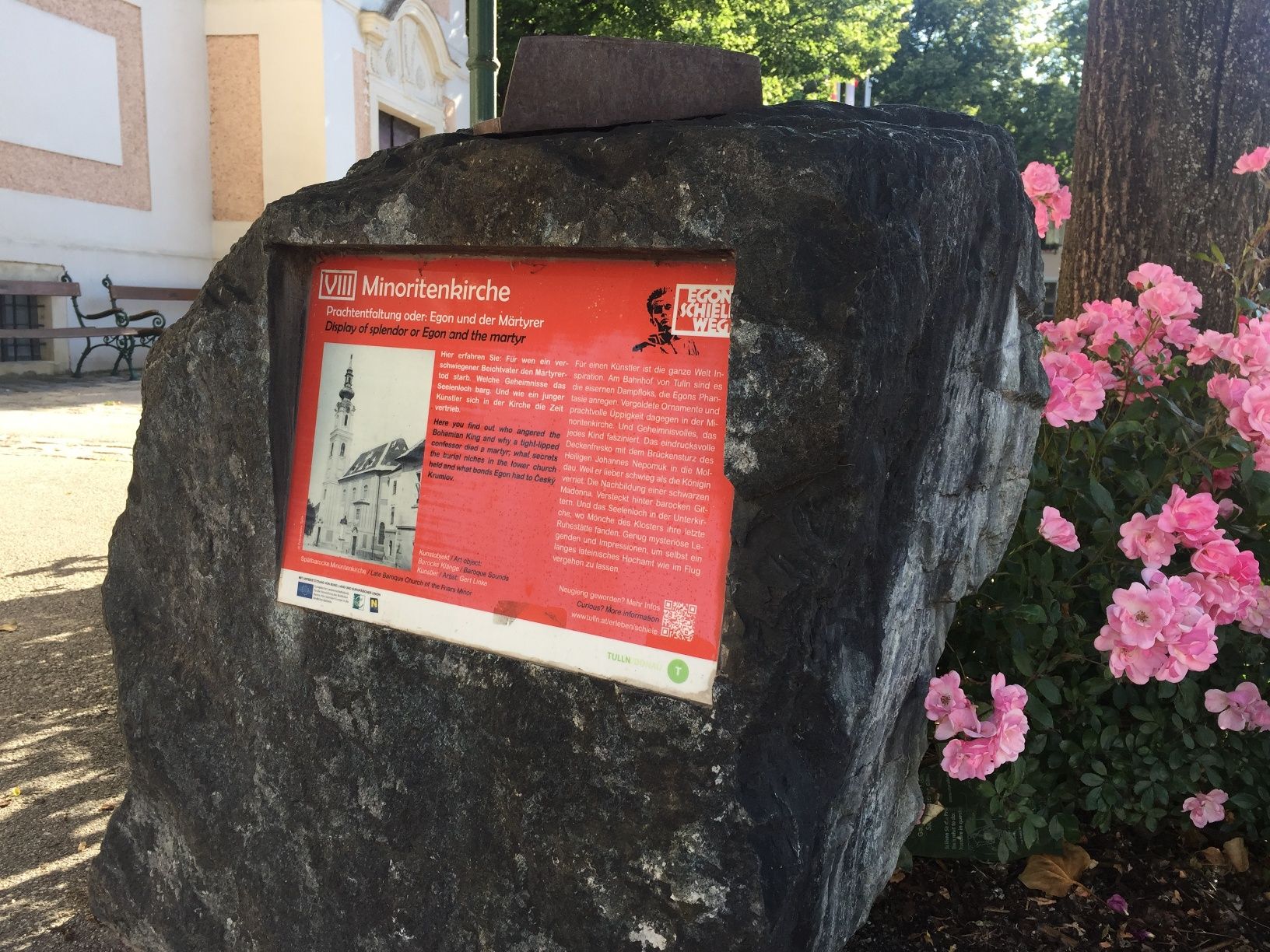 Kamenná tabuľa s informáciami o minoritskom kostole, obklopená kvetmi.