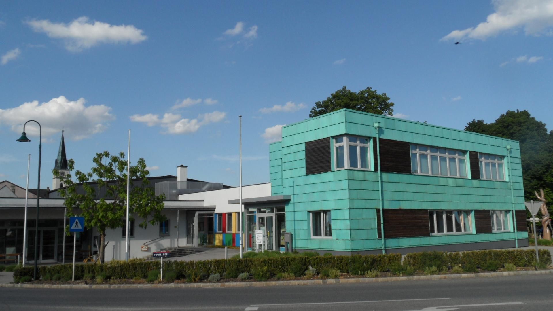 Moderná budova s tyrkysovo sfarbenou fasádou v Guntersdorfe, v pozadí kostolná veža.