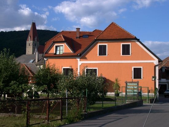 Dom oranžovej farby s červenými škridlami na streche a kostolnou vežou v pozadí.