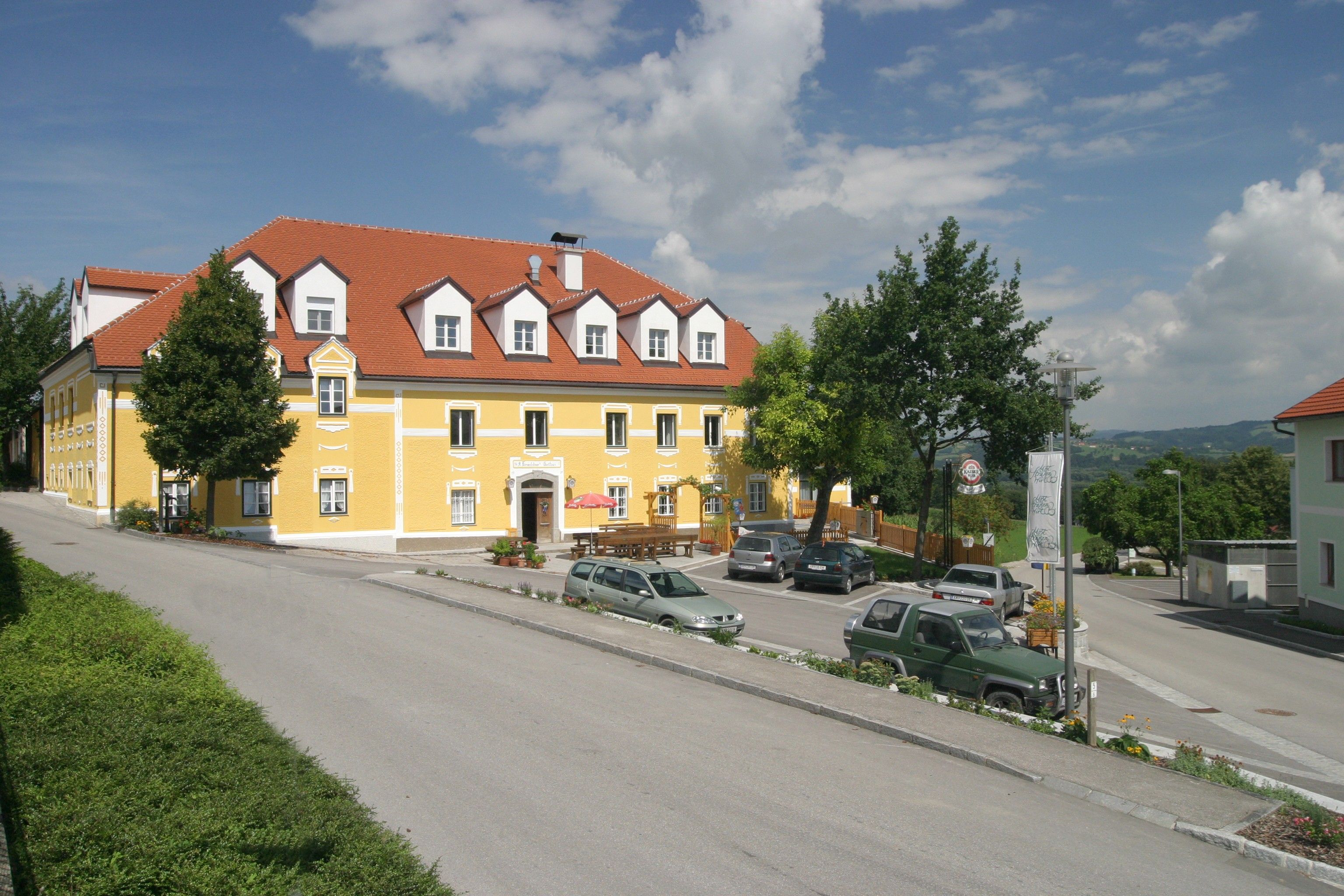 Žltá budova s červenou strechou, obklopená autami a stromami, pod modrou oblohou.