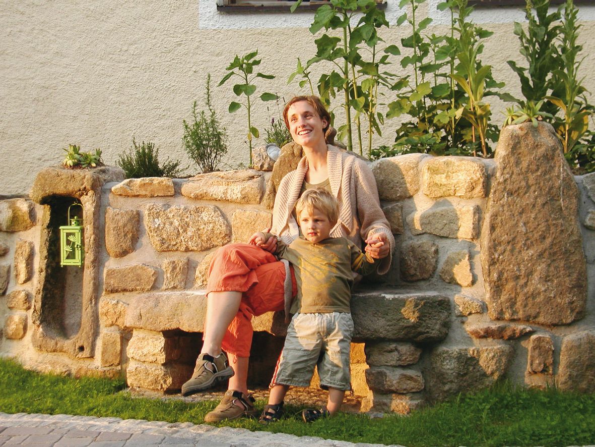 Žena a dieťa sedia na kamennej lavičke pred skalkou zarastenou rastlinami.