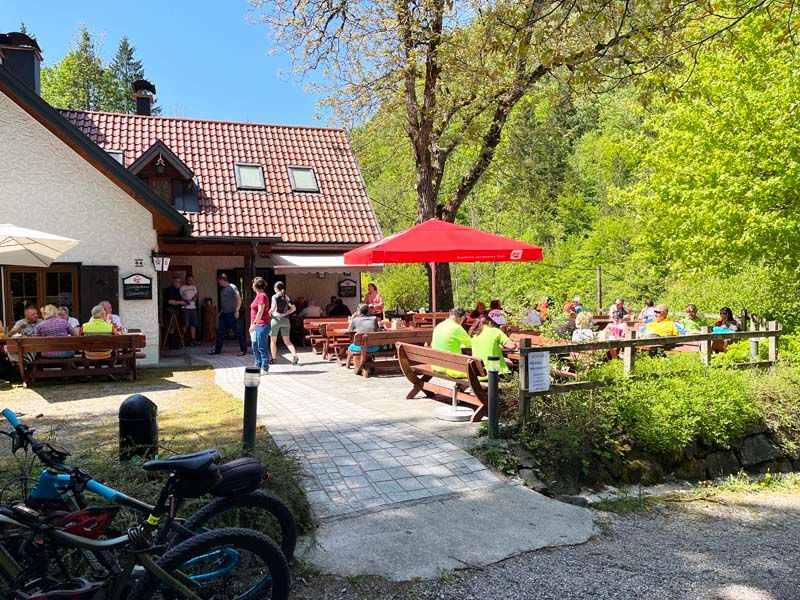Horská chata s terasou, kde ľudia sedia pri stoloch. V popredí vidieť bicykle obklopené zelenou prírodou.