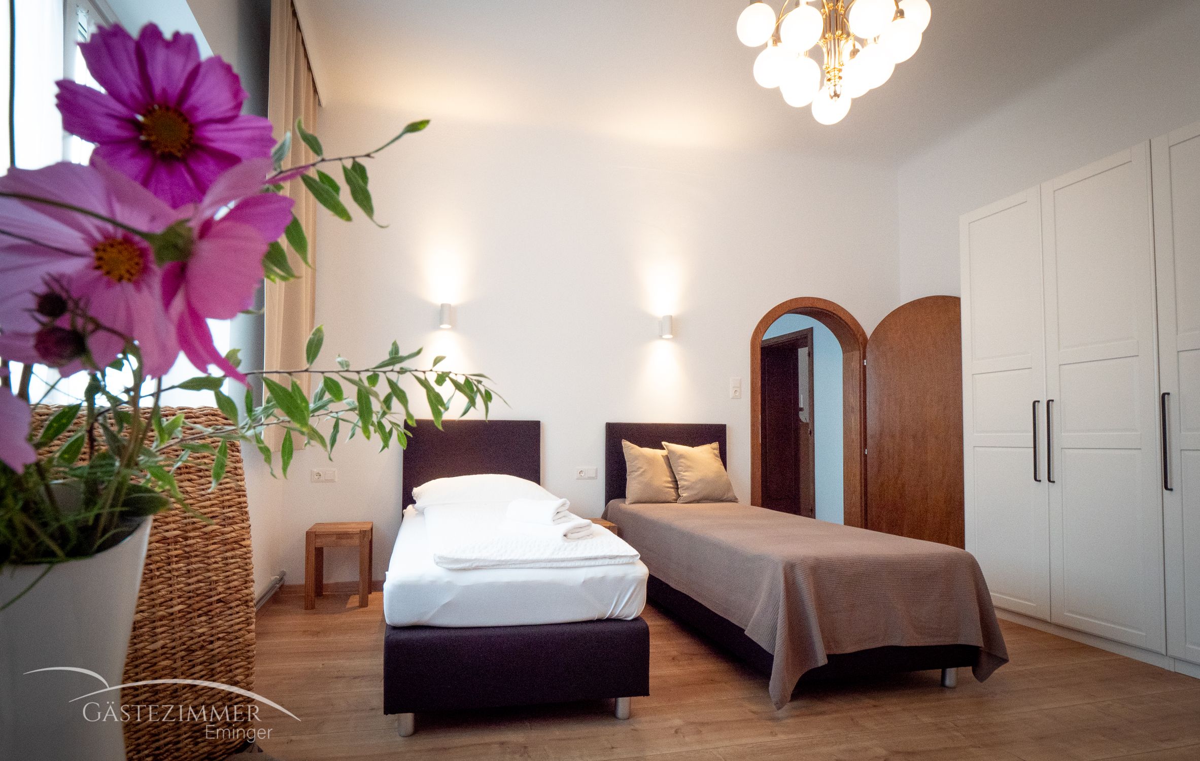 Útulná dvojlôžková izba s kvetmi v popredí, dvoma posteľami, šatníkovou skriňou a lampou na strope.