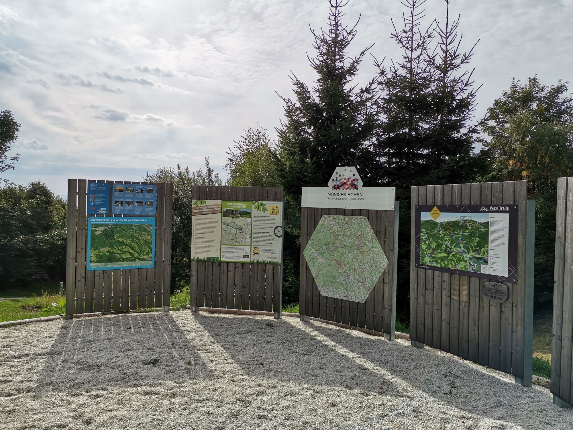 Informačné tabule na východiskovom bode turistiky v Mönichkirchene s mapami a informáciami, obklopené stromami.