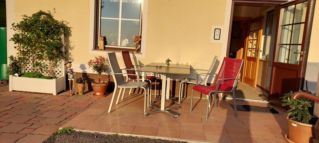 Terasa so stolom a stoličkami pred budovou, obklopená rastlinami.
