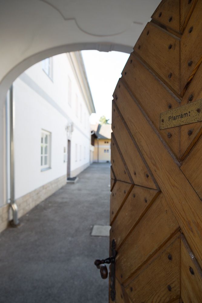 Drevená brána s nápisom "Pfarramt" sa otvára na nádvorie s bielymi budovami.