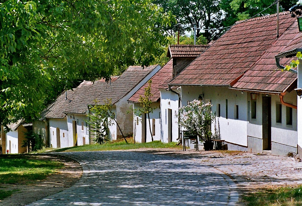 Dláždená ulica vedie okolo radu tradičných bielych domov s červenými strechami, obklopených zelenými stromami.