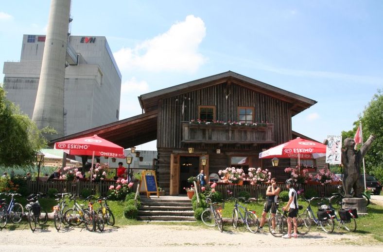 Drevená chatka s terasou, bicyklami a slnečníkmi pred priemyselnou budovou.