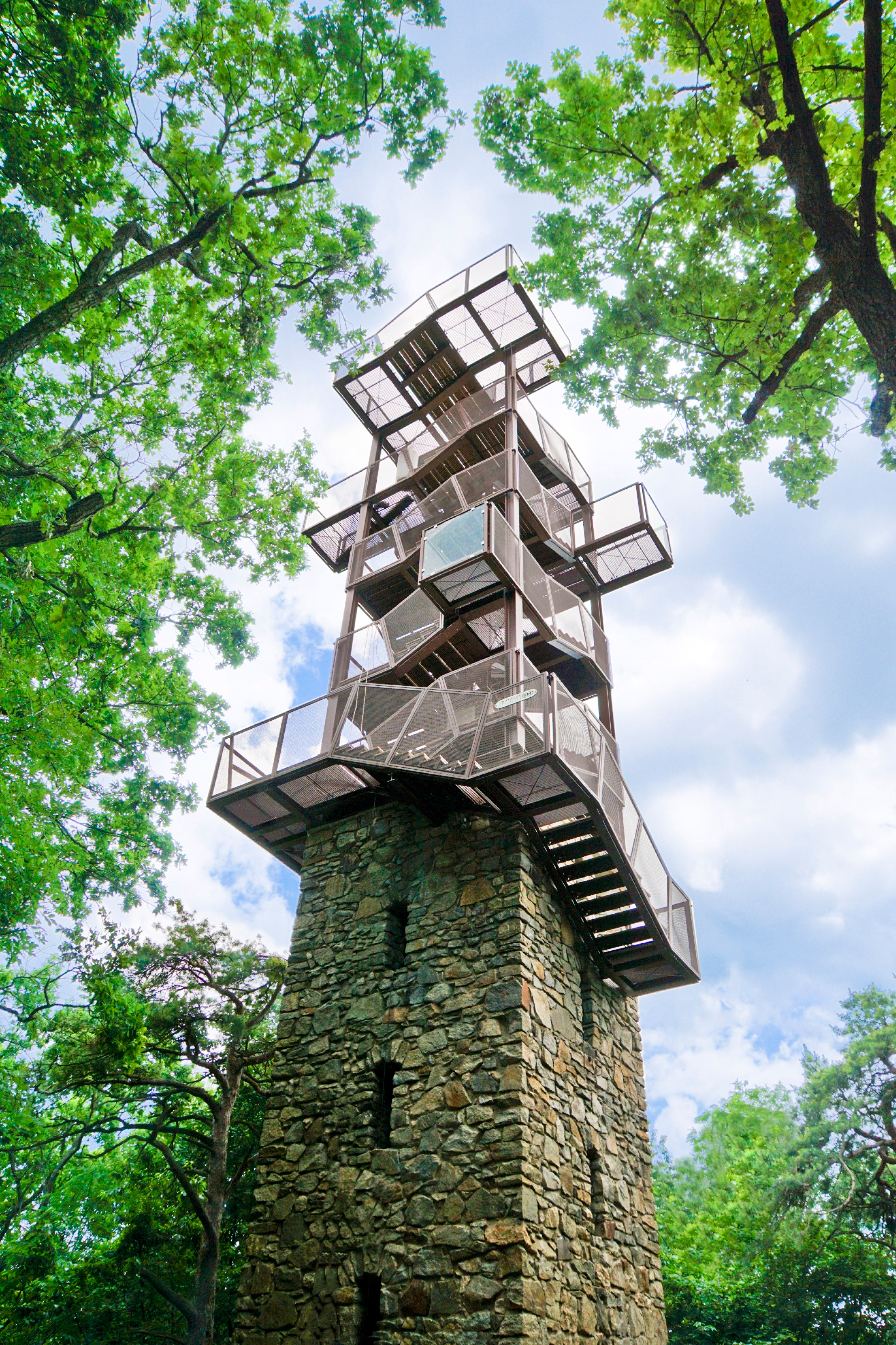 Do modrej oblohy sa týči moderná rozhľadňa z kovu a kameňa, obklopená zelenými stromami.