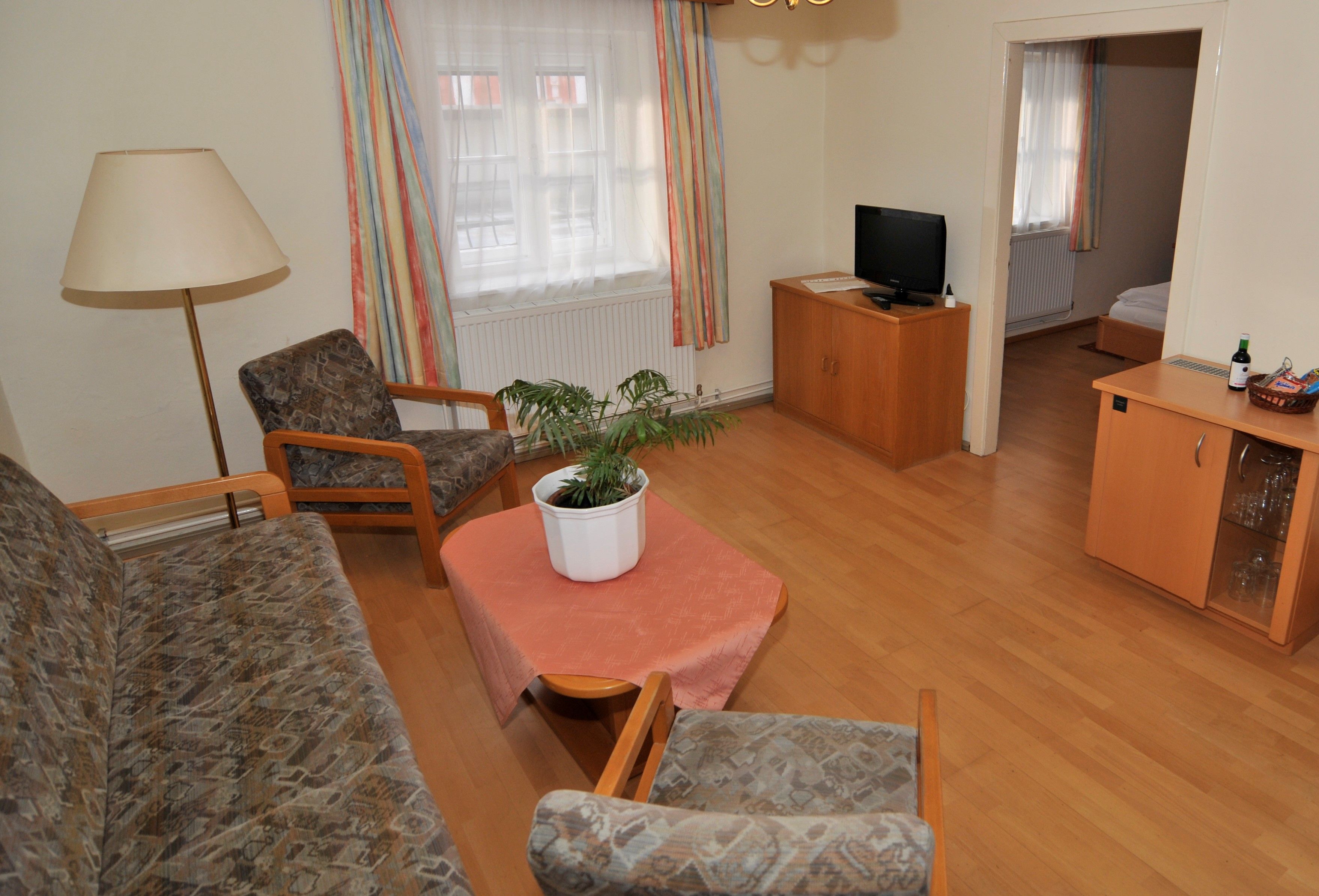 Obývacia izba s pohovkou, dvoma kreslami, stolom s rastlinami, TV a prístupom do spálne.