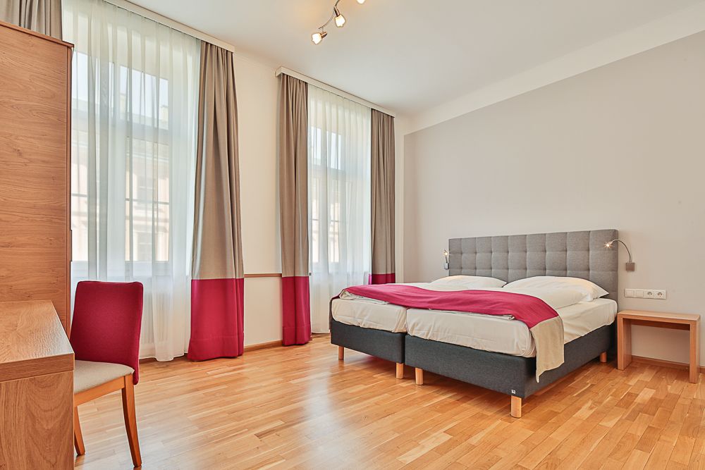 Svetlá hotelová izba s manželskou posteľou, stolom a stoličkou.