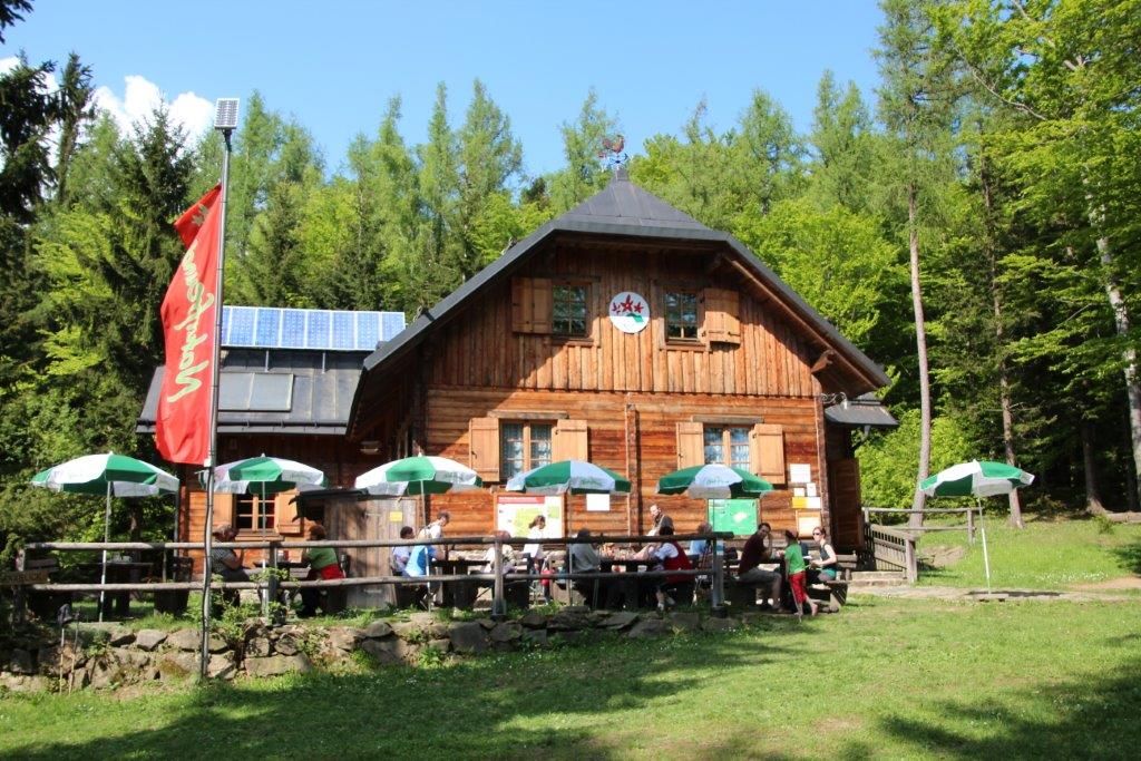 Drevená chata v lese so slnečníkmi a ľuďmi na terase.
