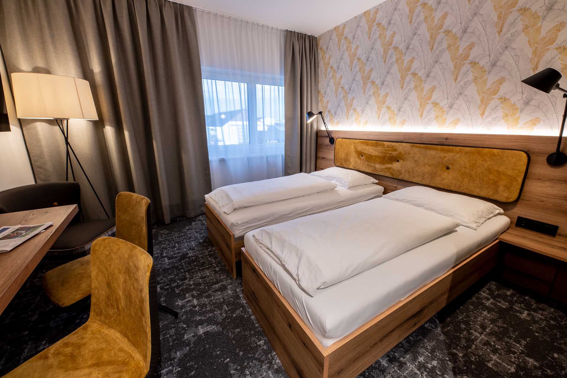 Moderná izba pre hostí s dvoma samostatnými posteľami, dreveným nábytkom, žltými stoličkami a stojacou lampou. Svetlé závesy a vzorovaná tapeta na stene.