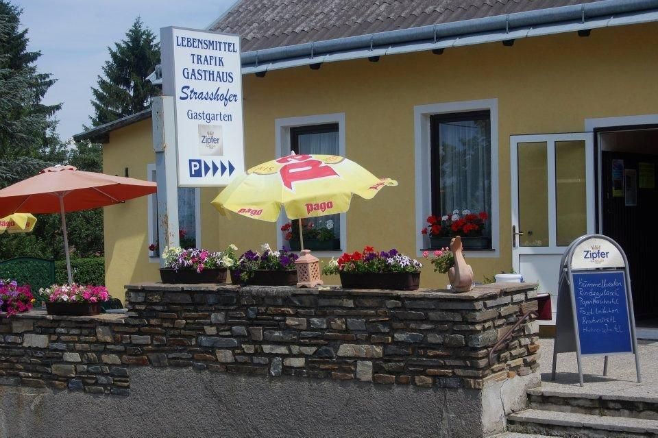 Pohľad zvonku na žltú budovu s nápismi na potraviny, trafiku a hostinec. Pred budovou sú slnečníky a kvety.