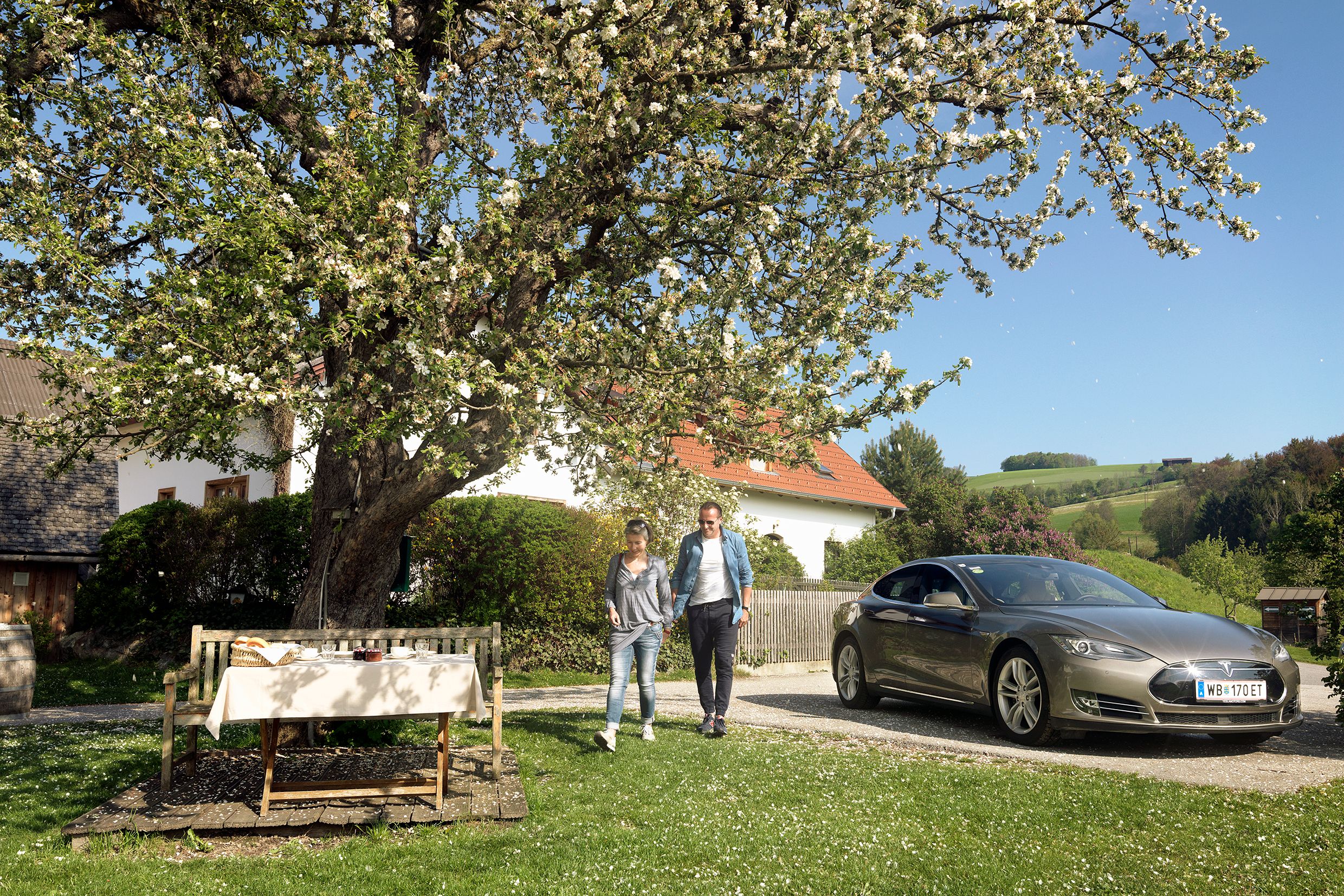 Pár kráča vedľa zaparkovaného elektromobilu pod rozkvitnutým stromom vo vidieckej záhrade.