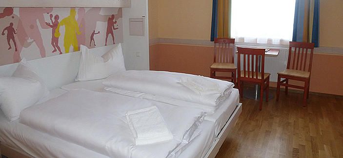 Hotelová izba s manželskou posteľou, dvoma stoličkami a stolom pred oknom.