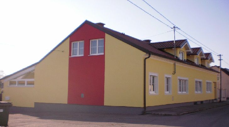 Pohľad zvonku na žlto-červenú budovu s niekoľkými oknami a šikmou strechou.