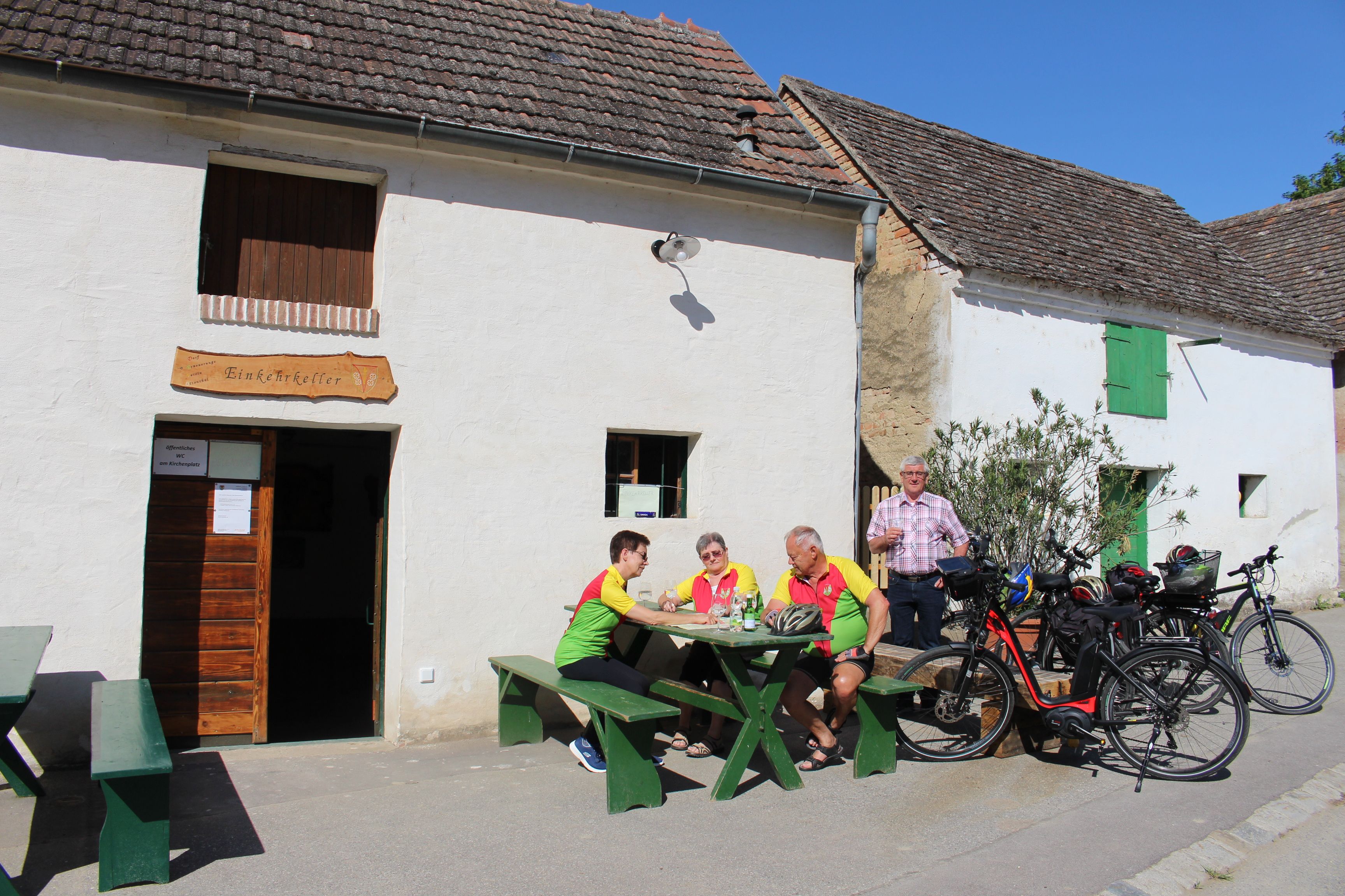 Skupina ľudí sedí pri stole pred reštauráciou „Einkehrkeller Ottenthal“. Vedľa sú zaparkované bicykle. Je slnečný deň.