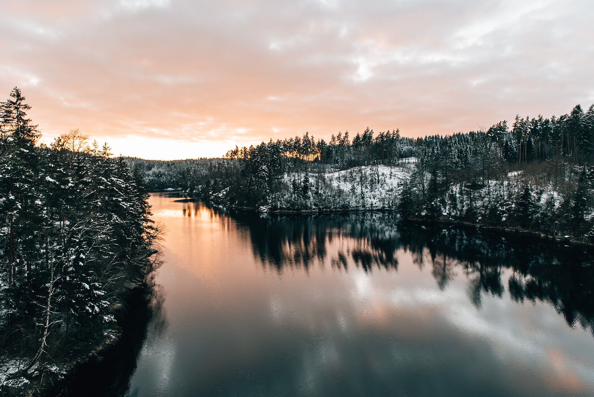 Jemné vlny nádrže odrážajú zasnežené brehy, zatiaľ čo zapadajúce slnko osvetľuje oblohu teplými farbami. Krajinu obklopuje pokojné ticho, ktoré vás pozýva na zotrvanie a oddych.