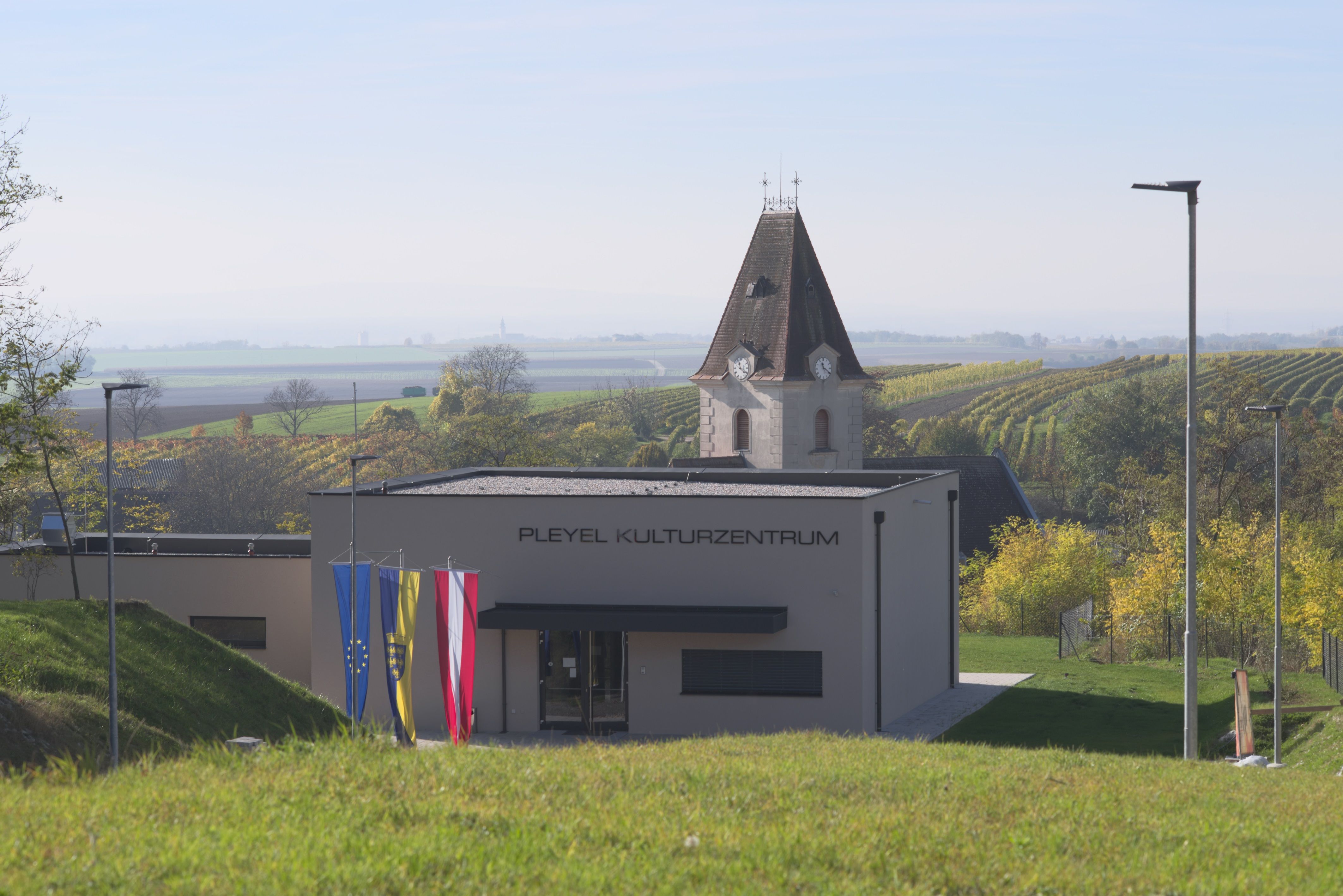Moderná budova s nápisom "Pleyel Cultural Centre" na pozadí vidieckej krajiny s vinicami a kostolnou vežou.