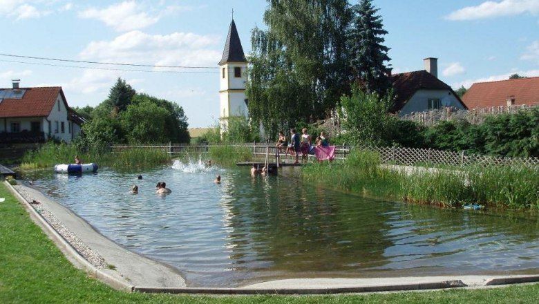 Prírodné kúpalisko s kúpajúcimi sa ľuďmi. V pozadí je vidieť kostolnú vežu a domy.