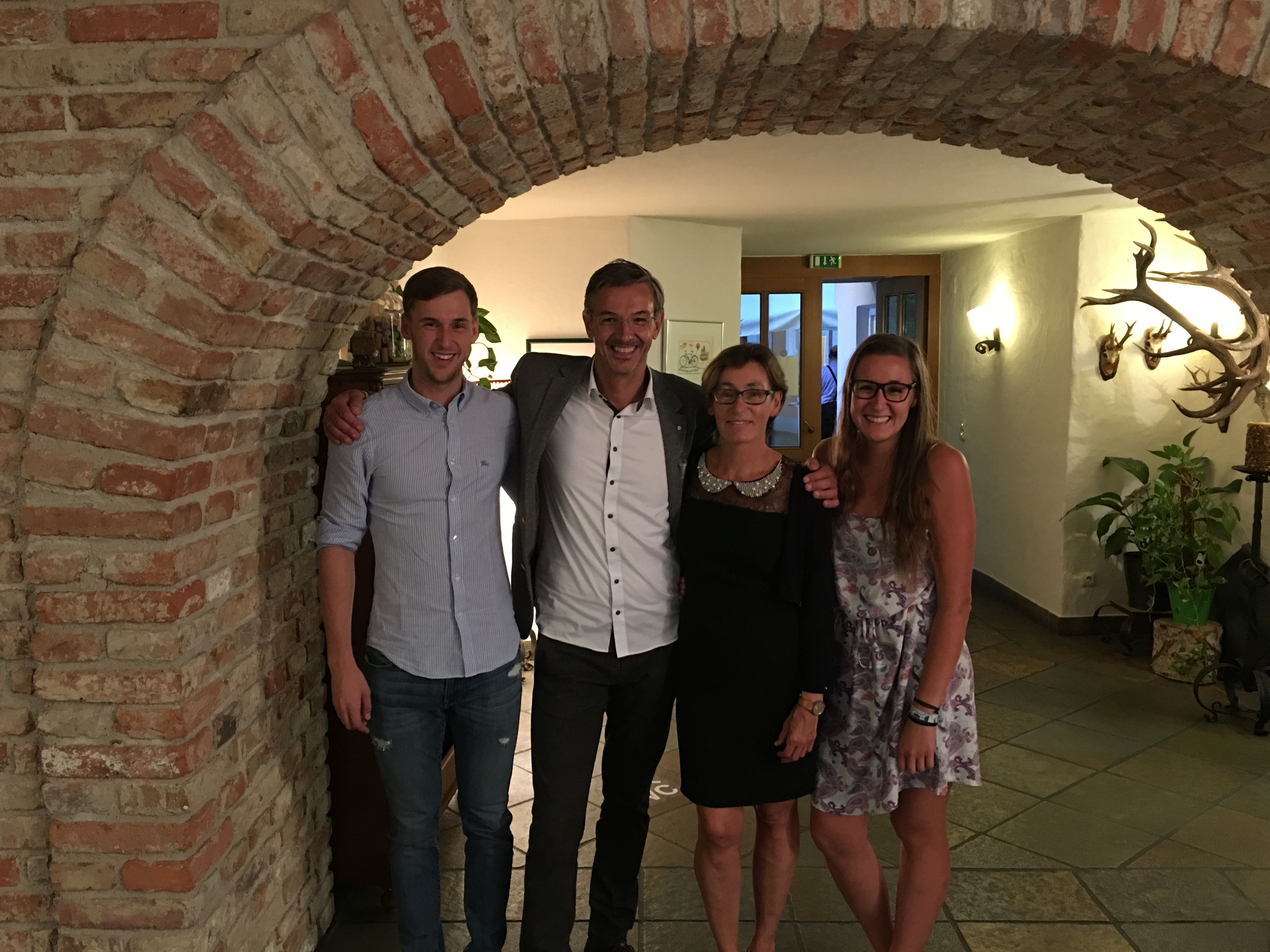 Štyria ľudia stoja s úsmevom pod tehlovým oblúkom v miestnosti s poľovníckymi trofejami na stene.