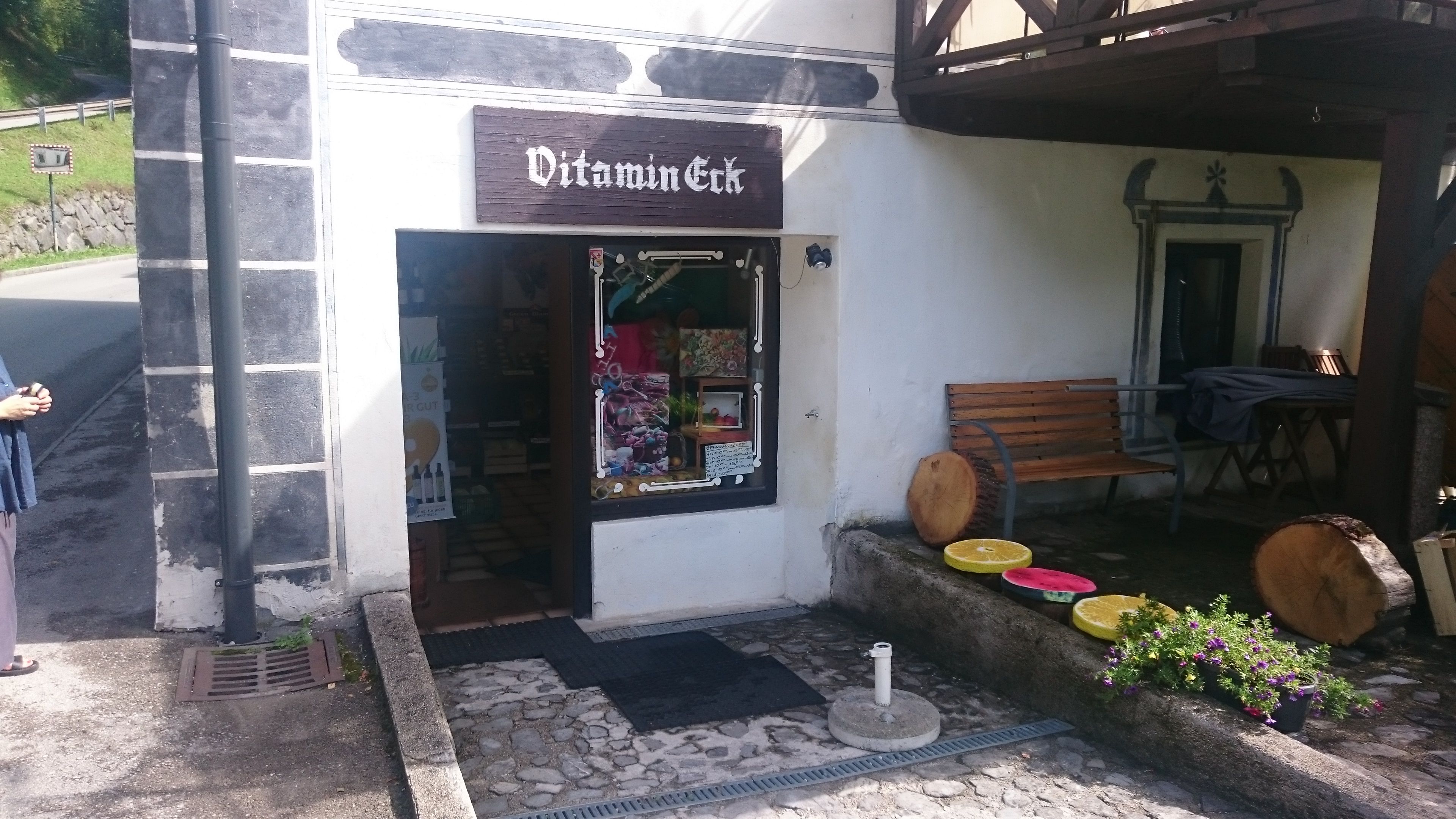 Vchod do malého obchodu Vitamin-Eck s lavičkou a dekoráciami z dreva a kvetov.