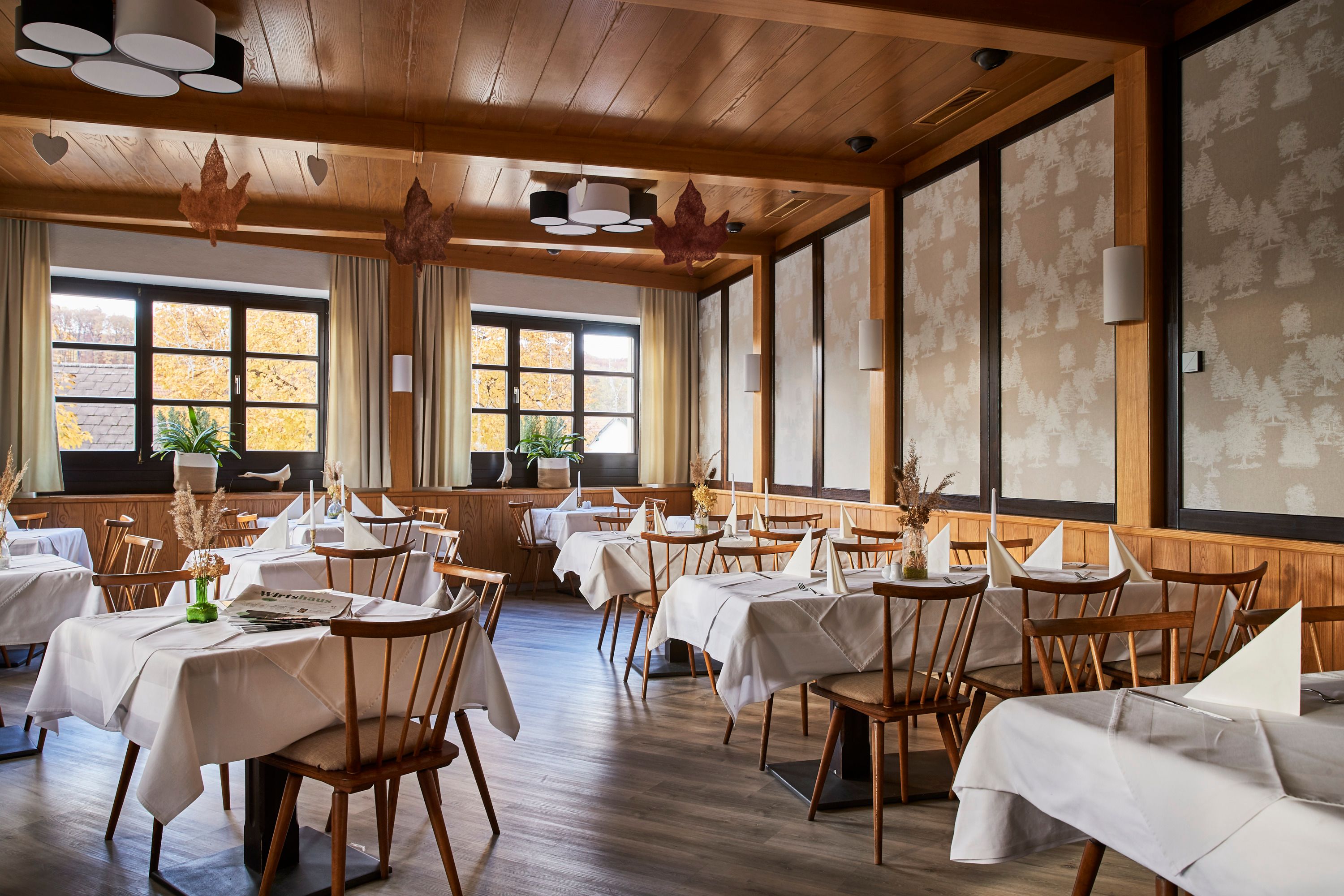 Elegantná reštaurácia s dreveným nábytkom, bielymi obrusmi a veľkými oknami s jesennými stromami.