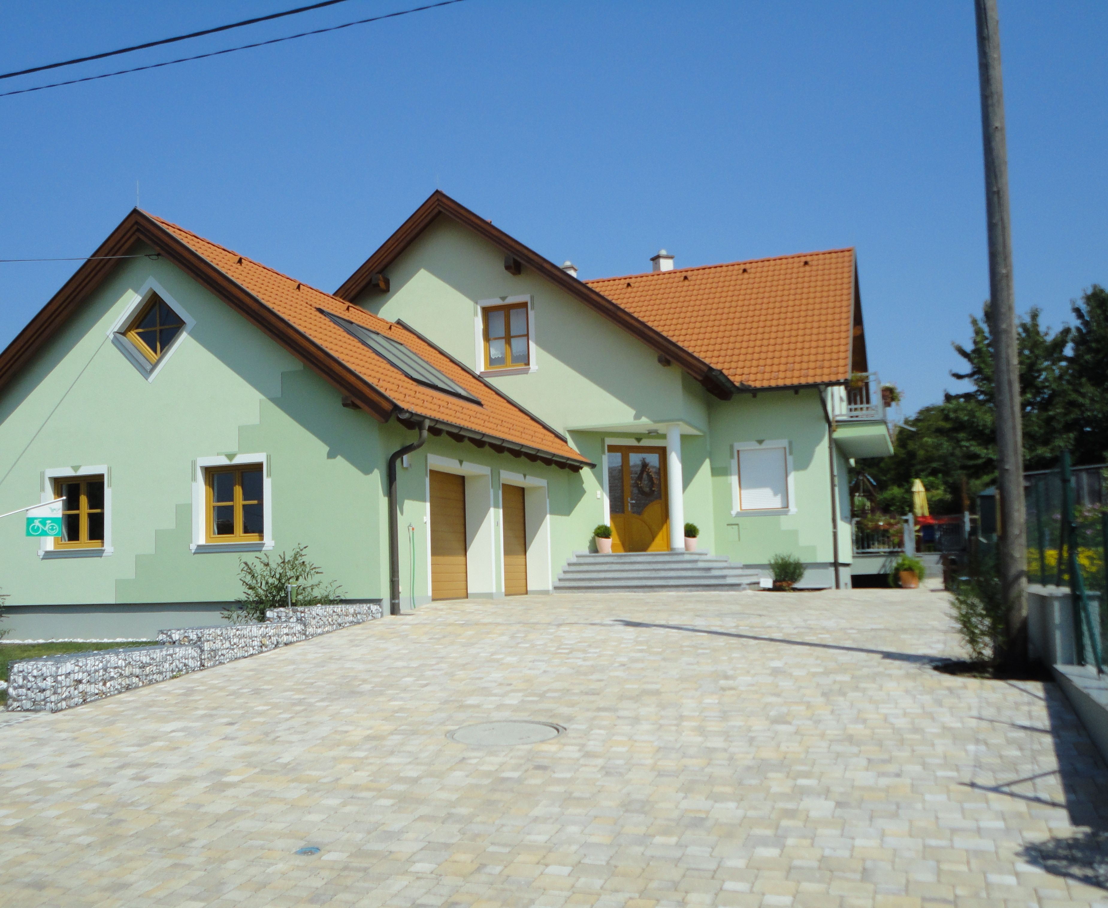 Moderný dvojpodlažný dom so zelenou farbou a červenými škridlami na streche, obklopený vydláždeným dvorom.