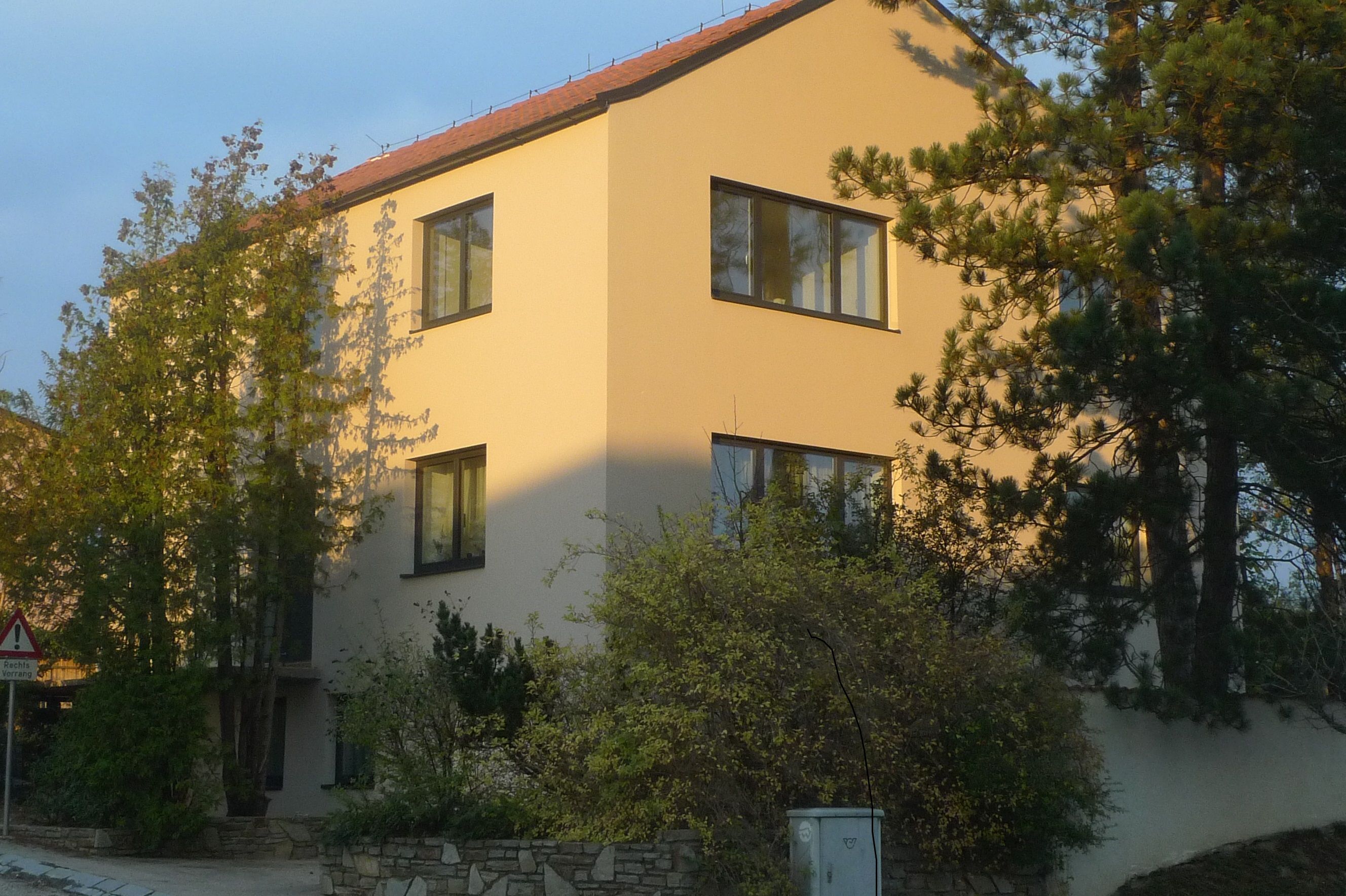 Moderný trojpodlažný dom so žltou fasádou a červenými škridlami, obklopený stromami a kríkmi.