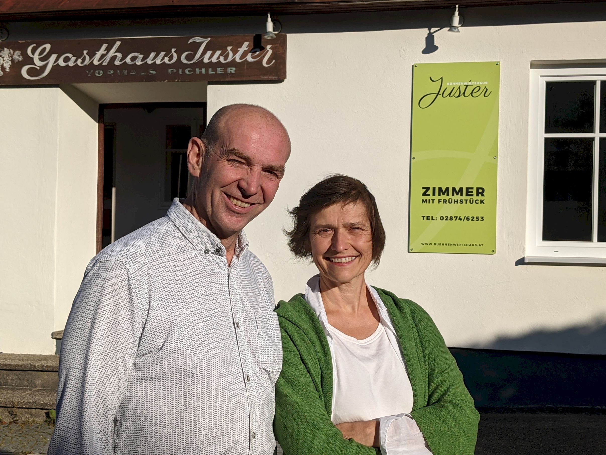 Muž a žena stoja s úsmevom pred hostincom Jüster. V pozadí je nápis "Izba s raňajkami".