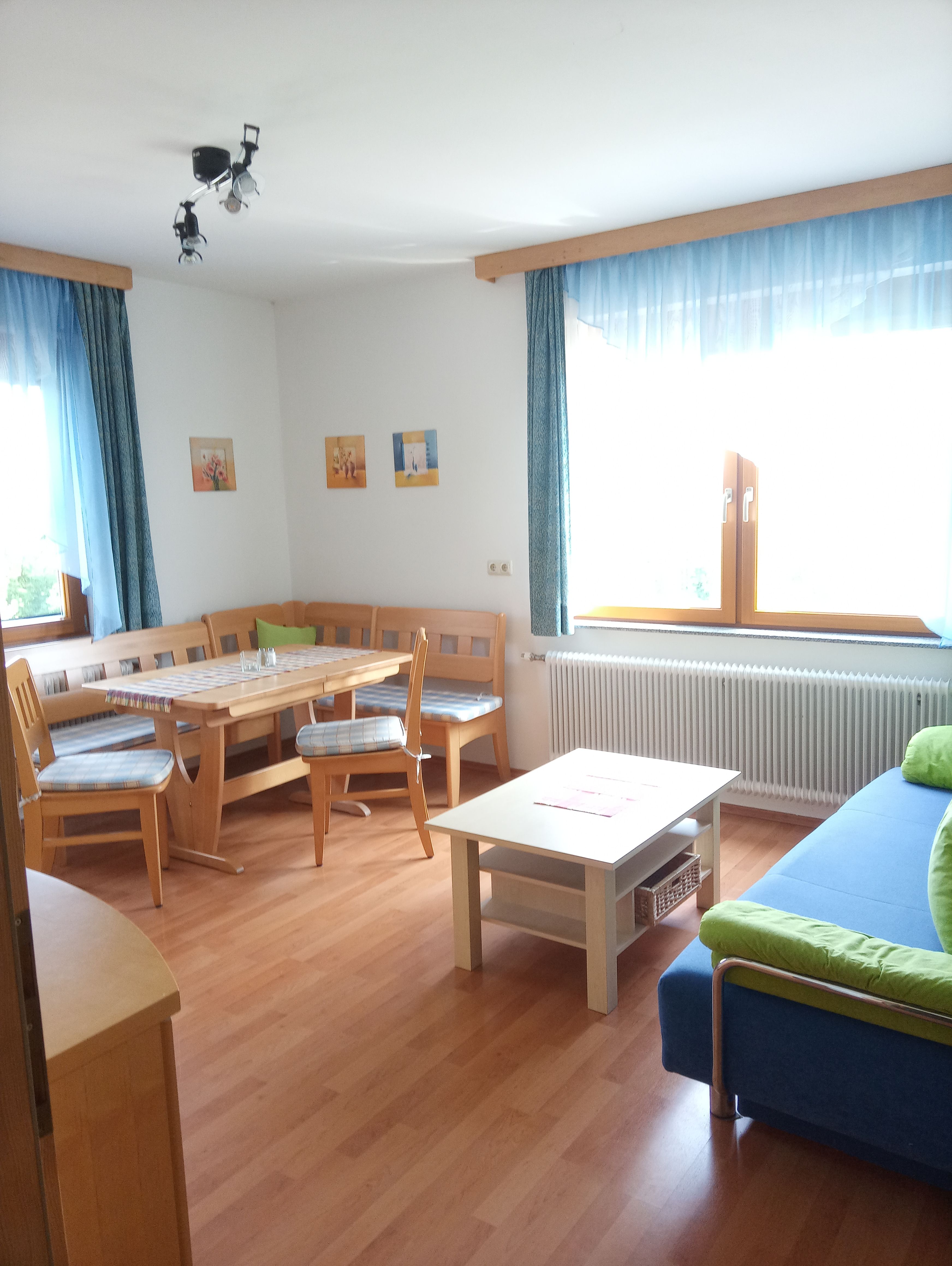 Svetlá obývacia izba s dreveným nábytkom, rohovou lavicou, stolom, pohovkou a veľkými oknami s modrými závesmi.