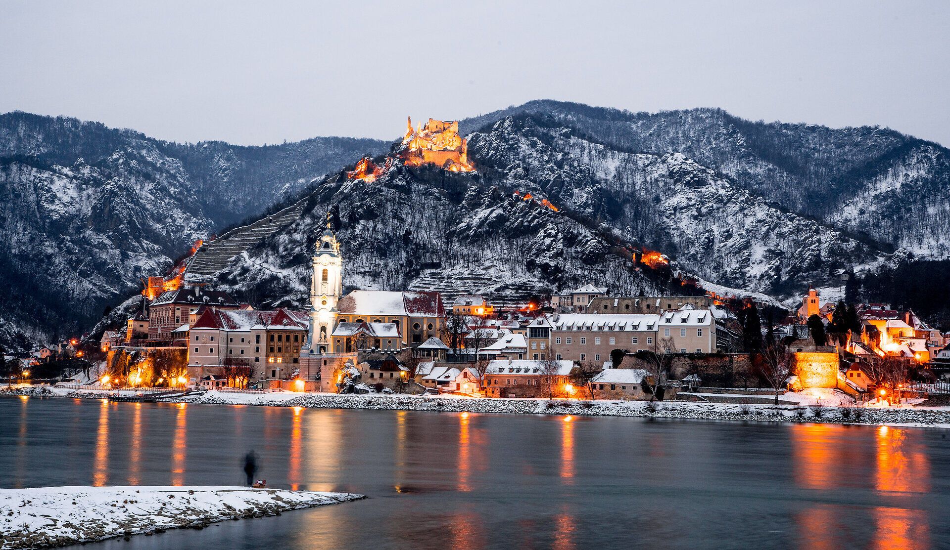 Die verschneite Landschaft um Dürnstein strahlt eine friedliche Ruhe aus, während die warmen Lichter der Stadt im Kontrast zur kalten Winterluft stehen. Die majestätische Ruine thront über dem blauen Wasser der Donau und lädt dazu ein, die winterliche Idylle zu erkunden.