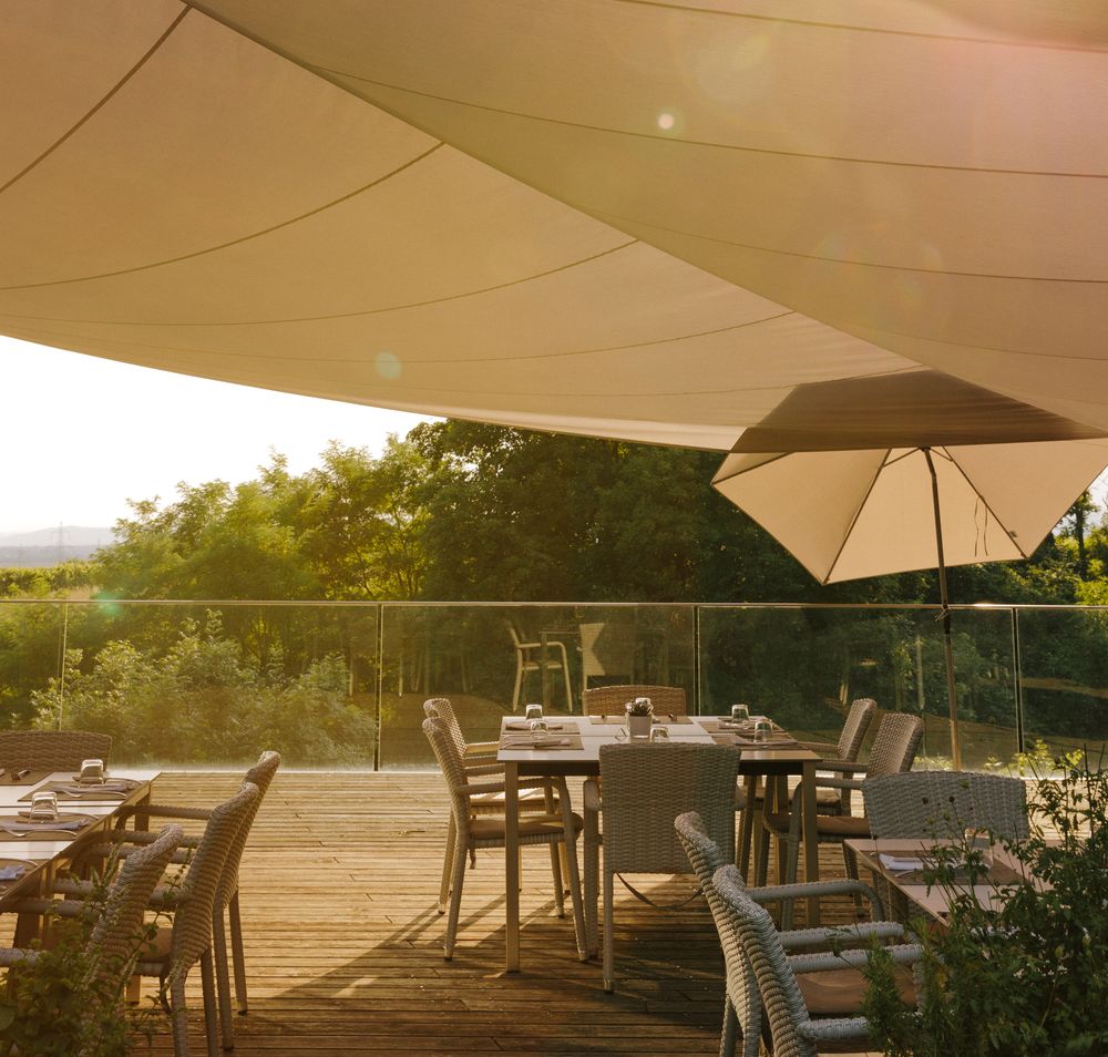 Unter einem sanften Sonnenschein laden die stilvollen Tische und Stühle auf der Terrasse zu einem entspannten Aufenthalt ein. Die sanften Farben des Himmels und die umgebende Natur schaffen eine harmonische Atmosphäre, die zum Verweilen einlädt. Hier kann man die Seele baumeln lassen und die Schönheit der Umgebung genießen.