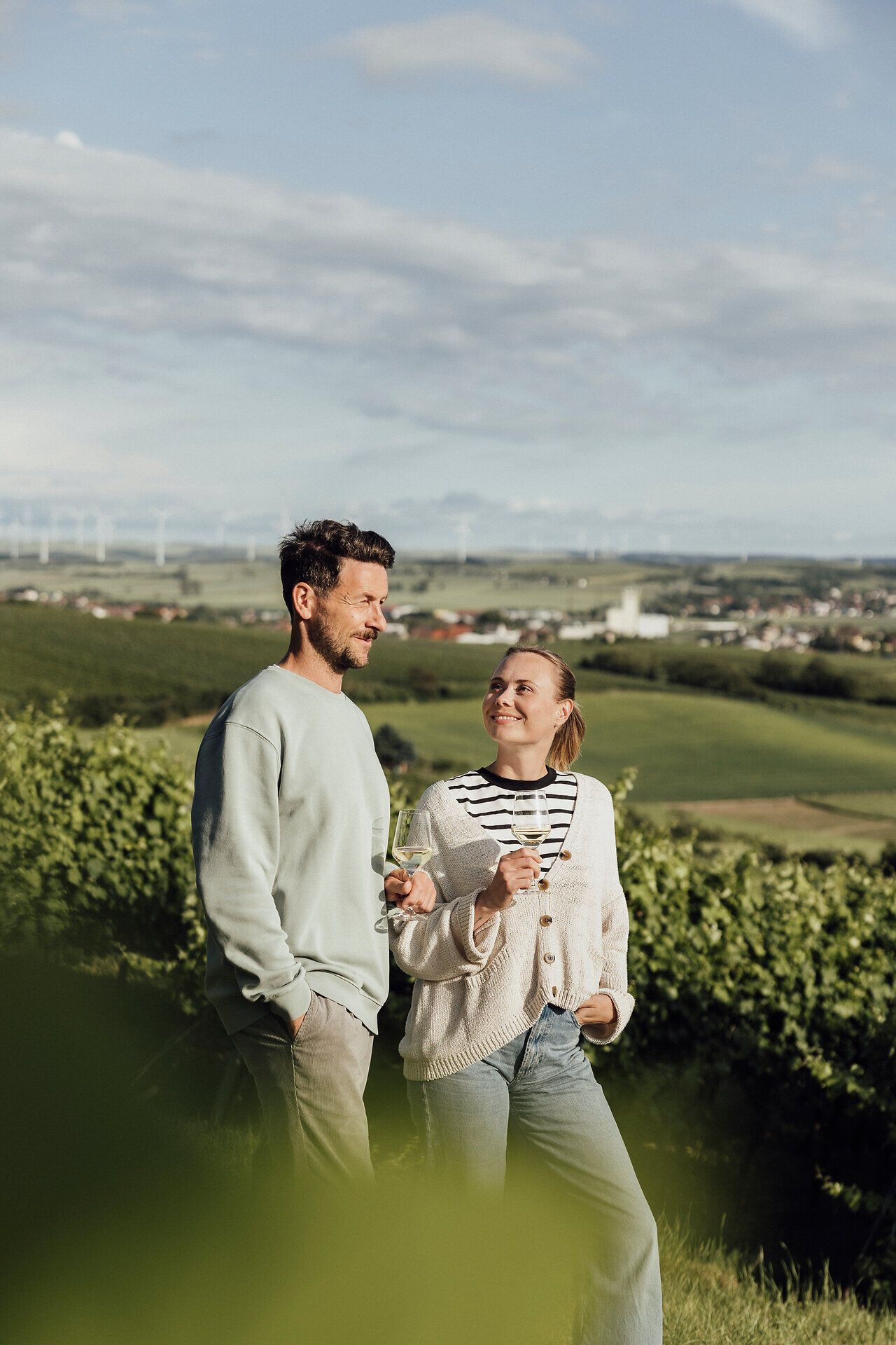 Inmitten der sanften Hügel des Weinviertels genießen zwei Menschen ein Glas Wein und lassen sich von der malerischen Landschaft verzaubern. Die grüne Umgebung und der weite Blick laden dazu ein, die Seele baumeln zu lassen und den Moment in vollen Zügen auszukosten.