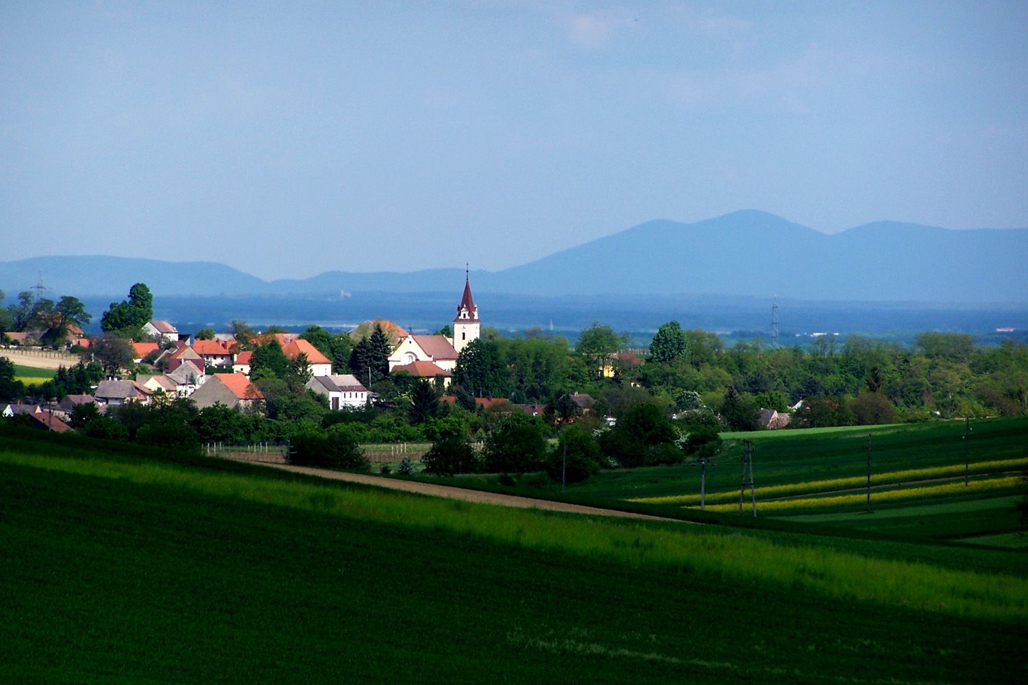 Krajina s dedinou a kostolom, obklopená zelenými poliami a kopcami v pozadí.