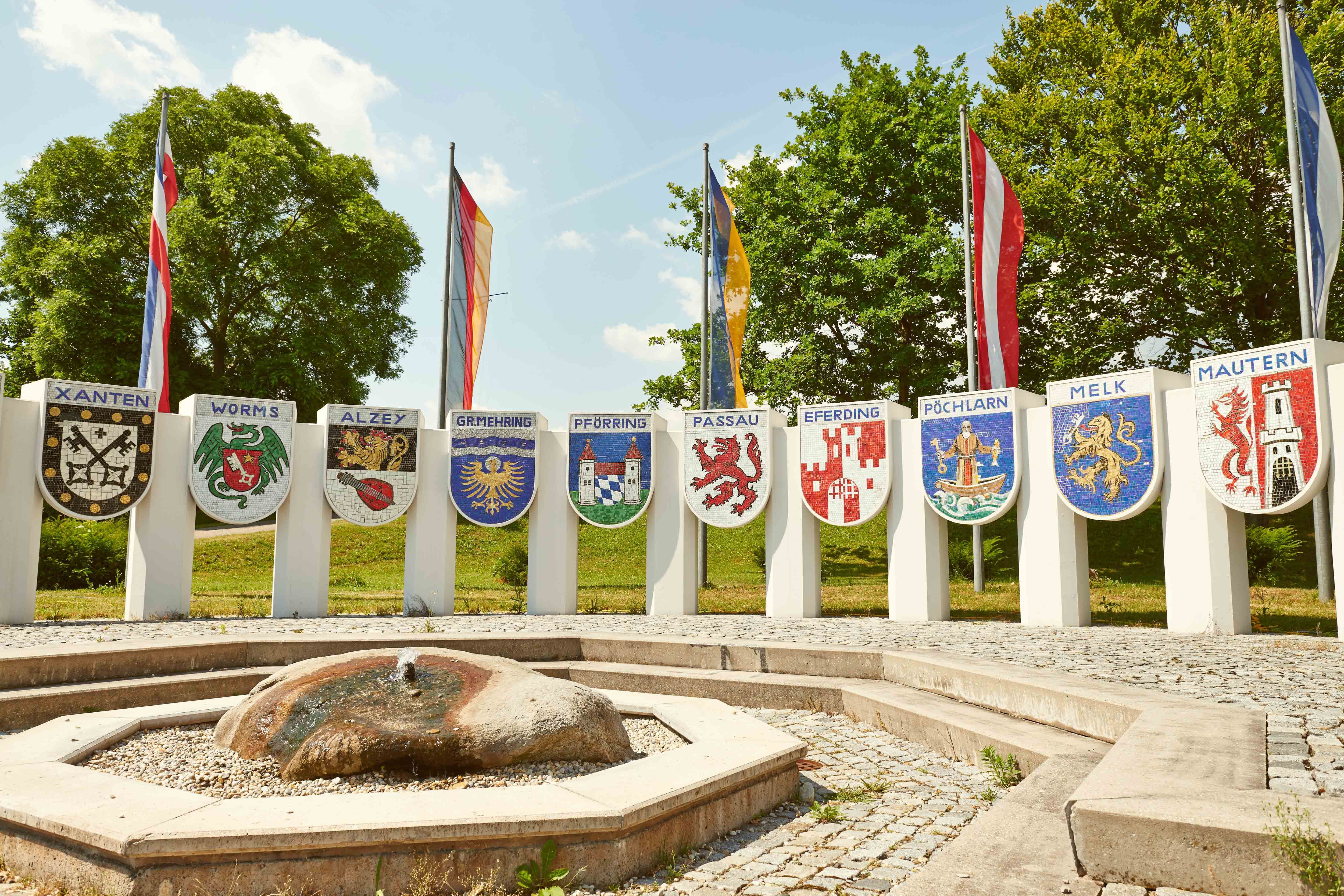Pomník Nibelungov s erbami rôznych miest a vlajkami v pozadí.