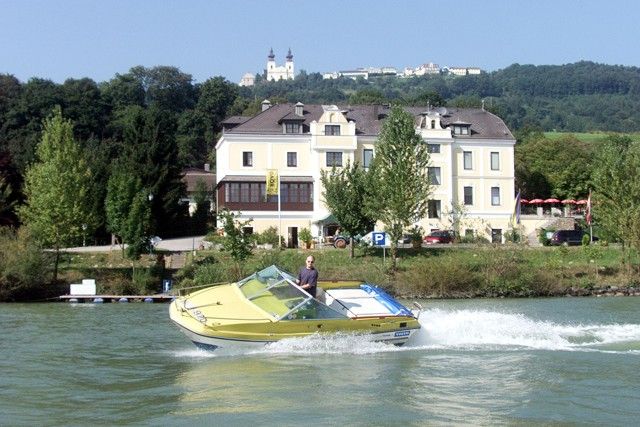 Žltý motorový čln sa plaví po rieke pred veľkou svetlou budovou s kopcami v pozadí.