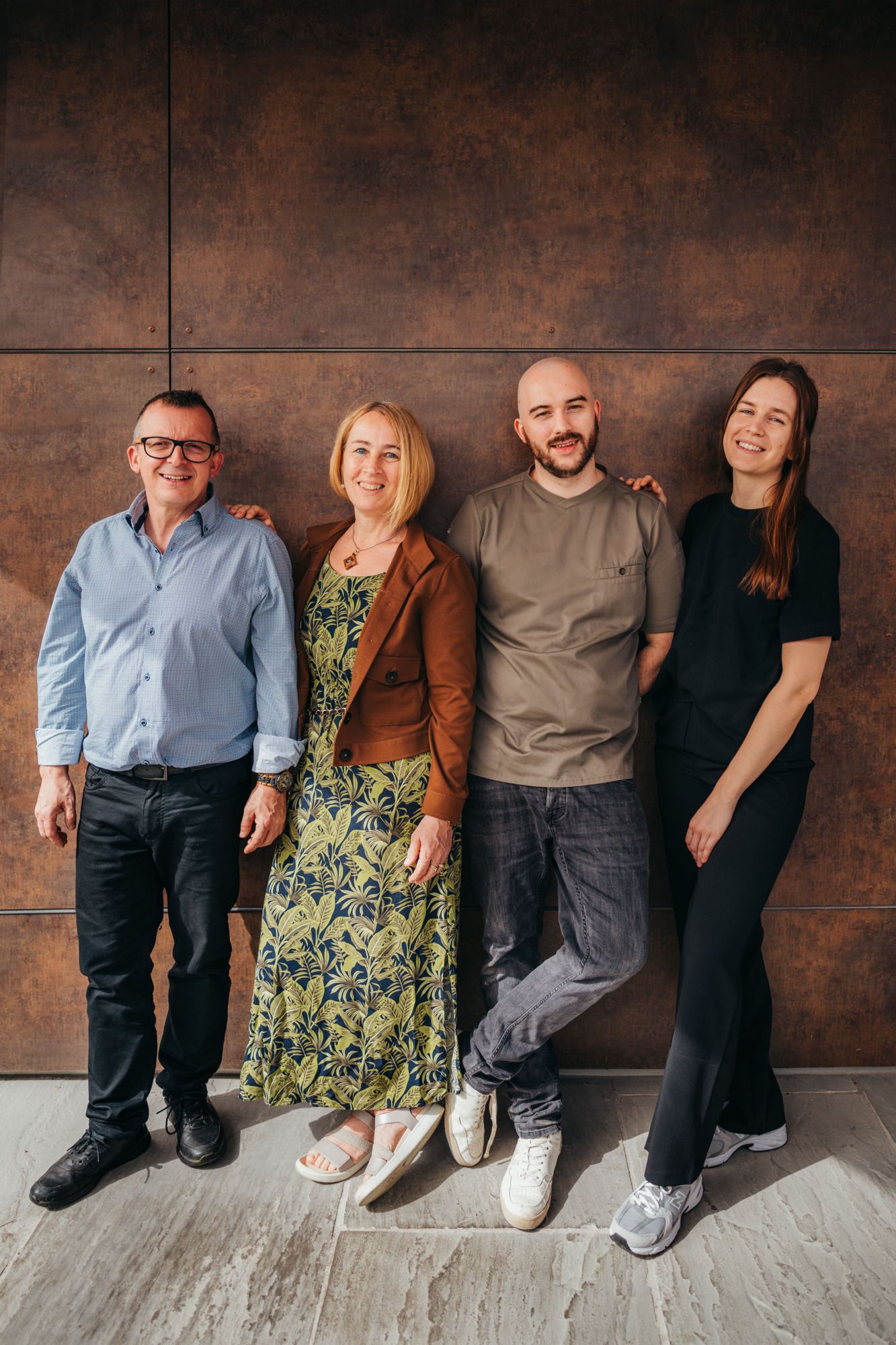 Štyria ľudia stoja s úsmevom vedľa seba pred hnedou stenou.