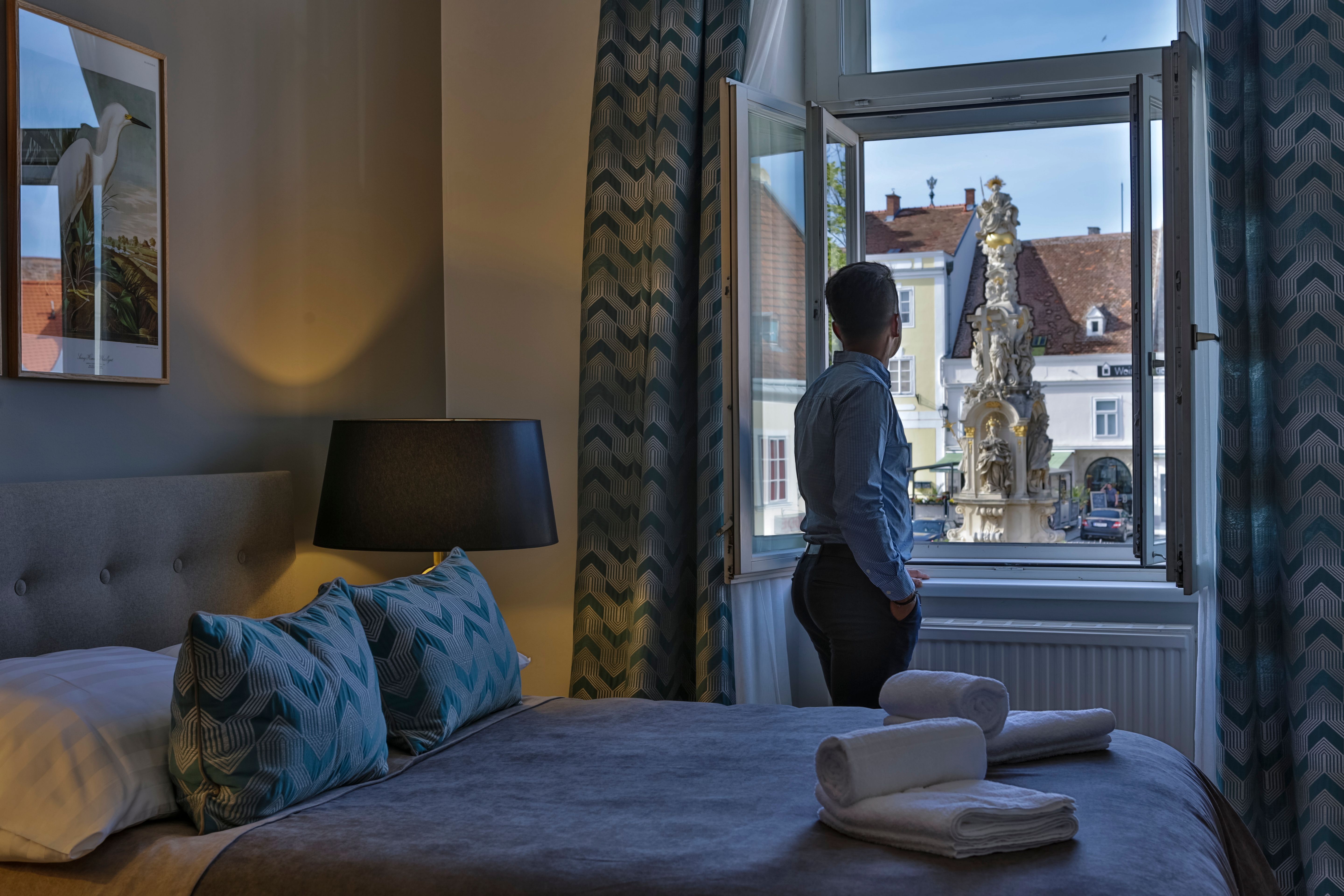 Osoba stojí pri okne hotelovej izby a pozerá sa na fontánu vonku.