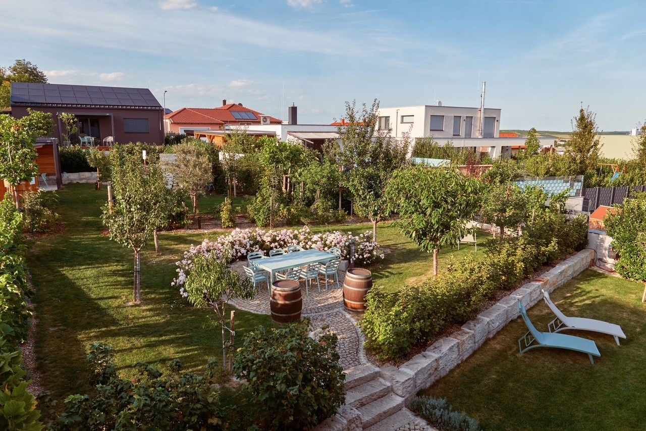 Dobre udržiavaná záhrada so stromami, kvetmi, stolom so stoličkami a dvoma ležadlami. V pozadí vidieť moderné domy.