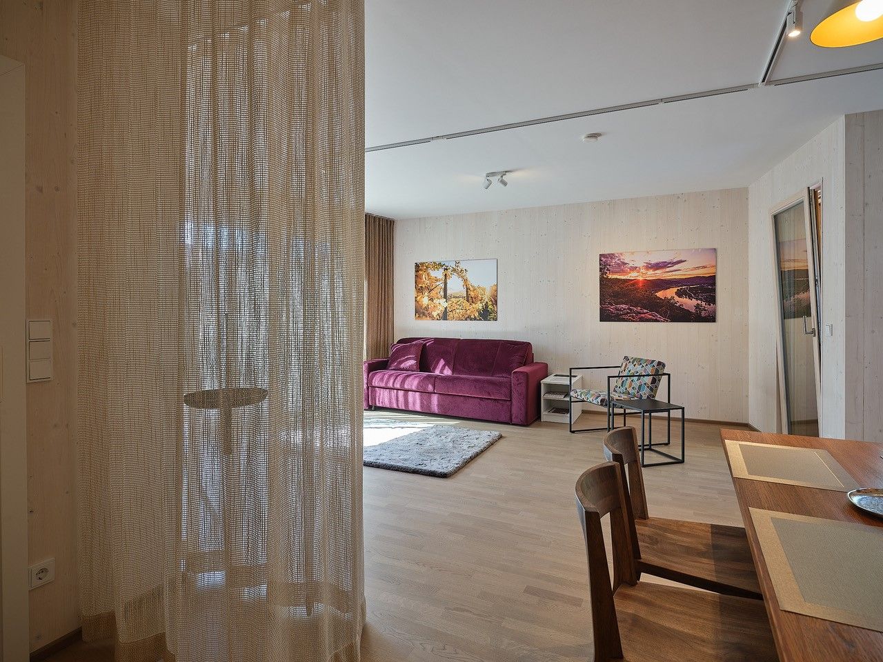 Obývacia izba s fialovou pohovkou, dreveným stolom a stoličkami, závesom a nástennými maľbami.