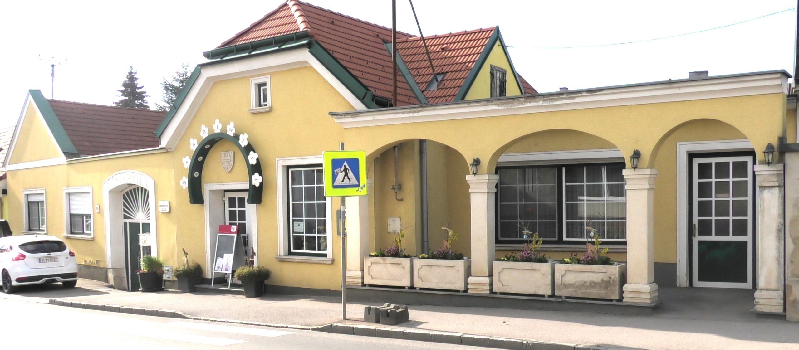 Pohľad zvonka na žltú budovu s arkádami a červenou strechou.