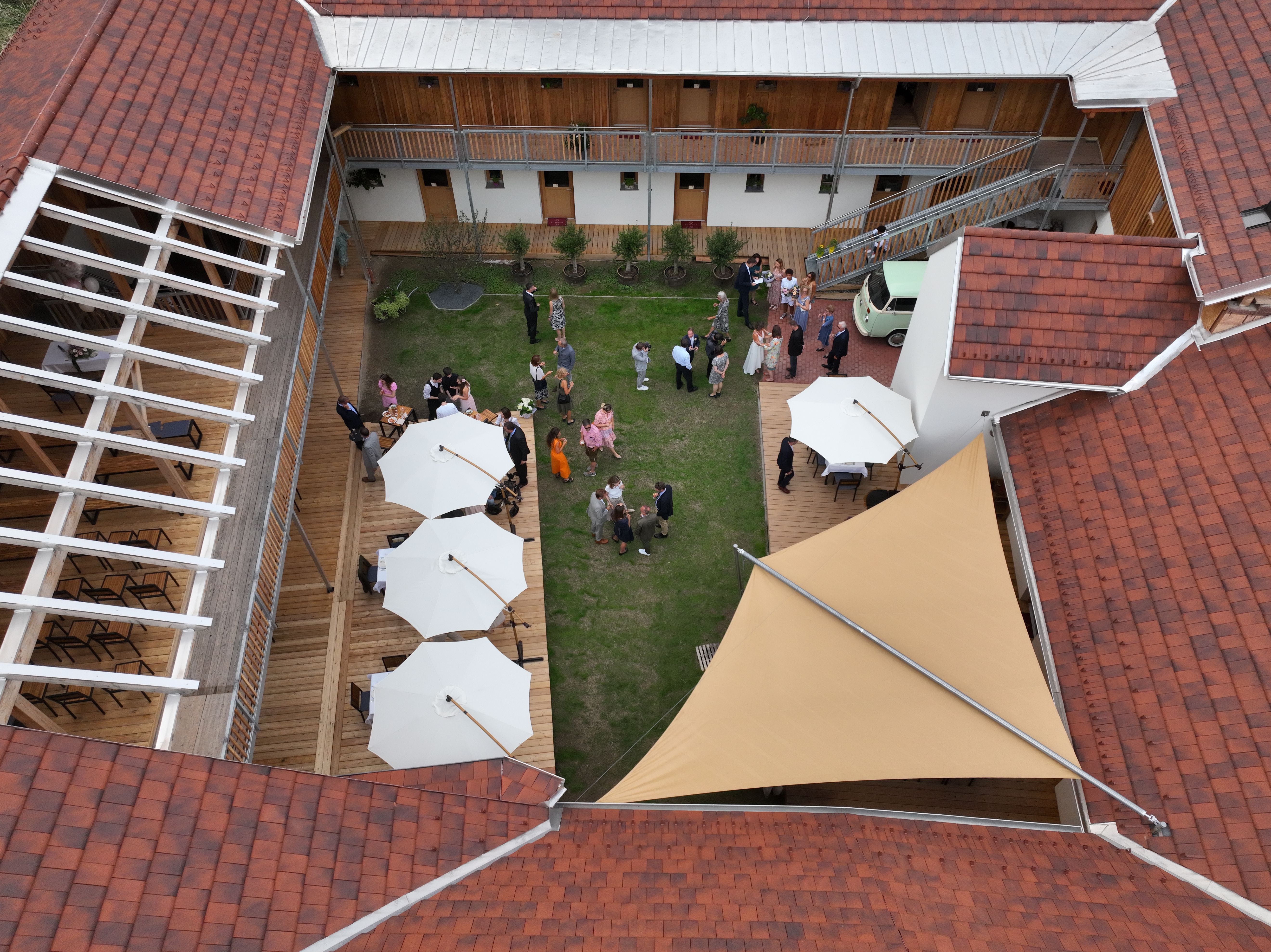 Pohľad z vtáčej perspektívy na vnútorné nádvorie so zeleným trávnikom, bielymi slnečníkmi a hosťami na recepcii.