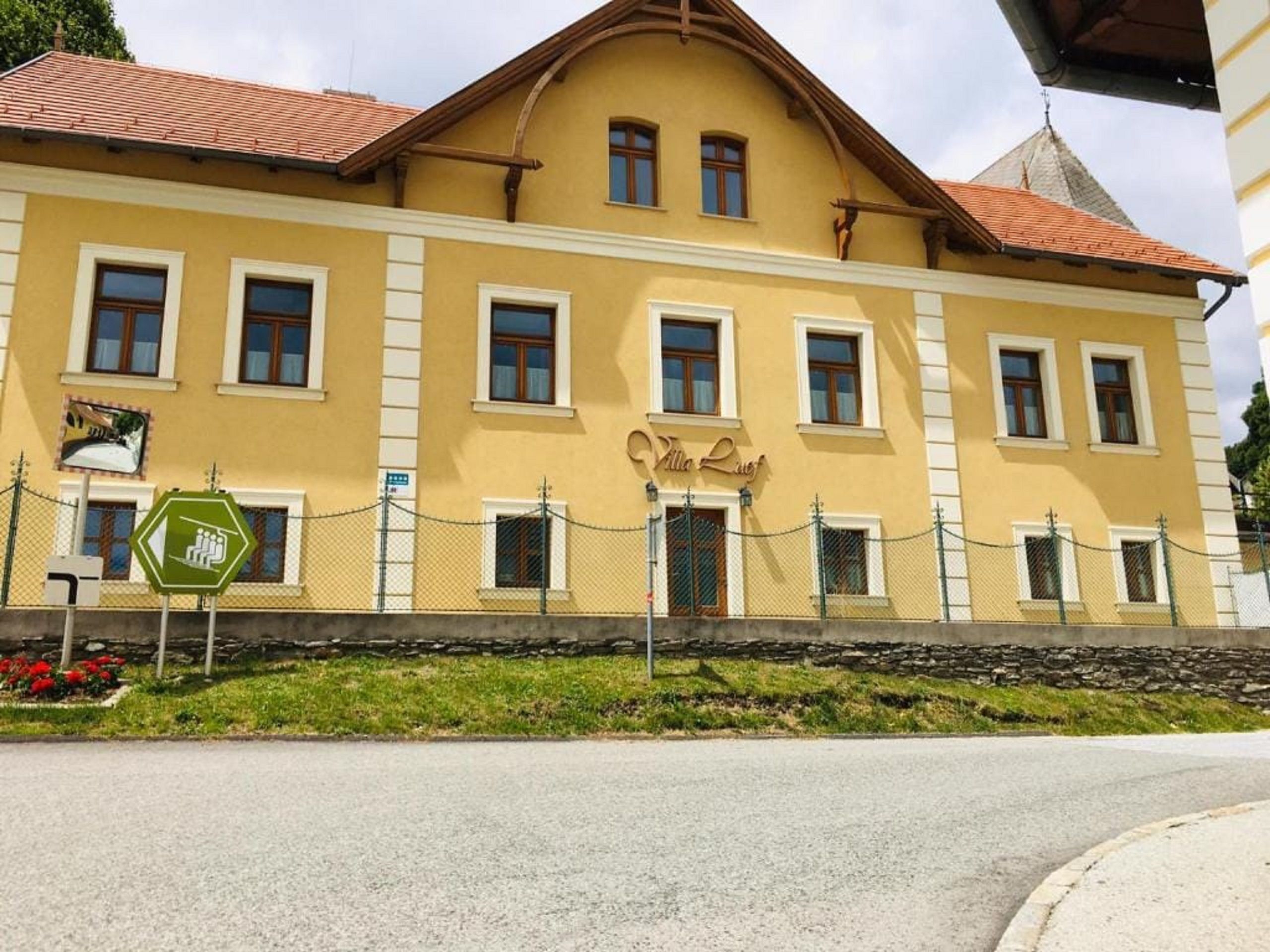 Žltá budova s nápisom "Villa Luef", obklopená plotom a cestou v popredí.